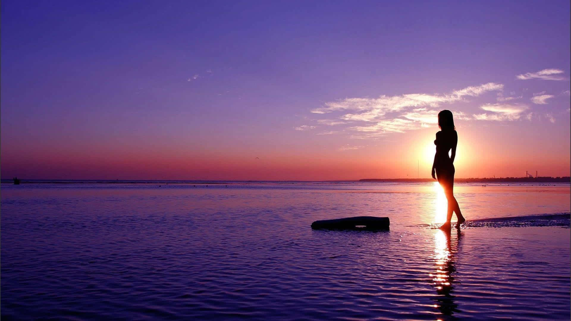 Sunset Silhouette 桌布 壁紙 — 日落時分，一個孤獨女性的剪影站在淺水中，她的倒影在平靜的水面上閃爍，背景是紫色和橙色的漸變天空。