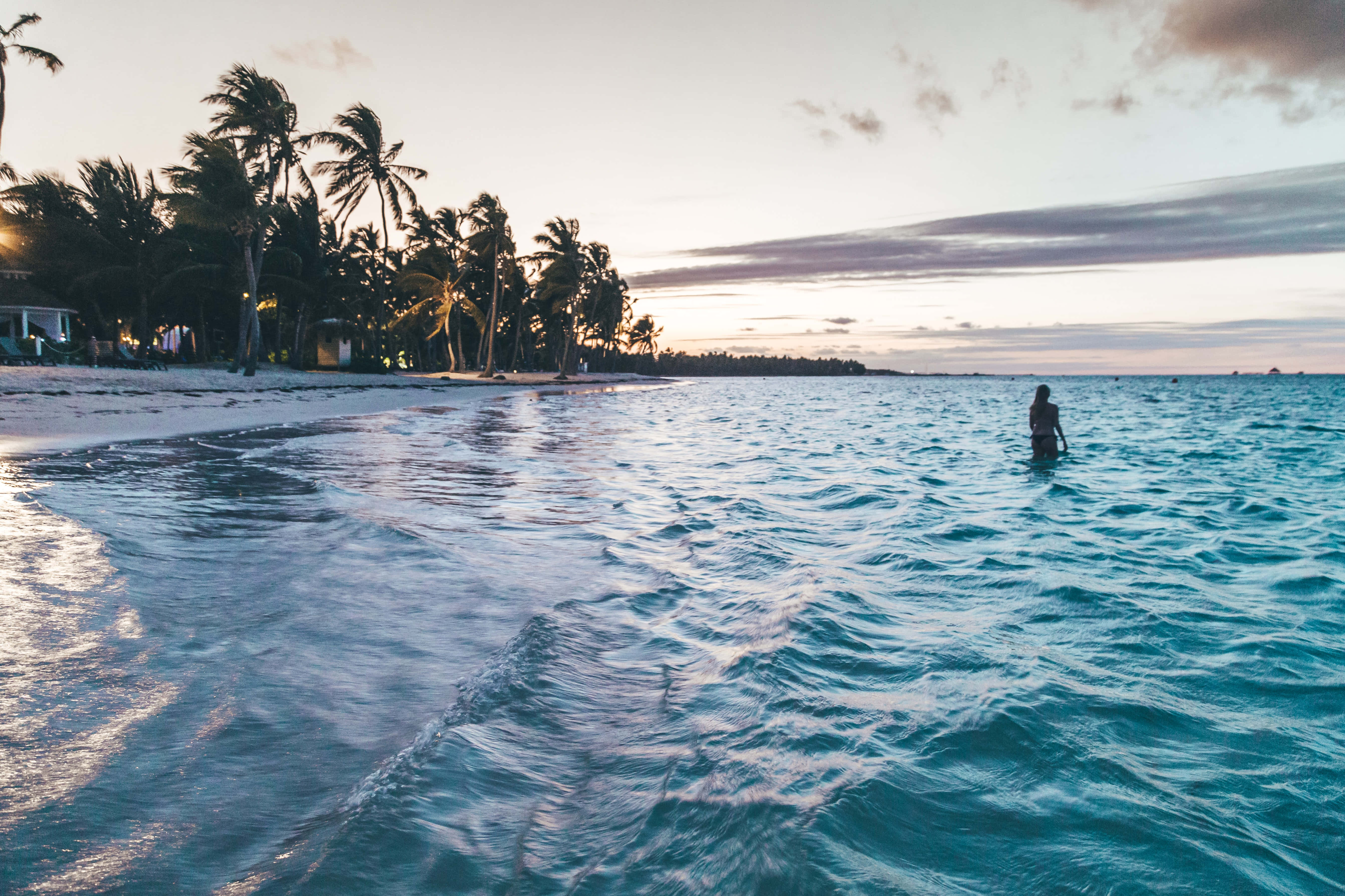 Caribbean Beach 桌布 壁紙 — 日落時分，一個孤獨的身影在碧綠的海水中涉水而行，沙灘上棕櫚樹的剪影清晰可見。