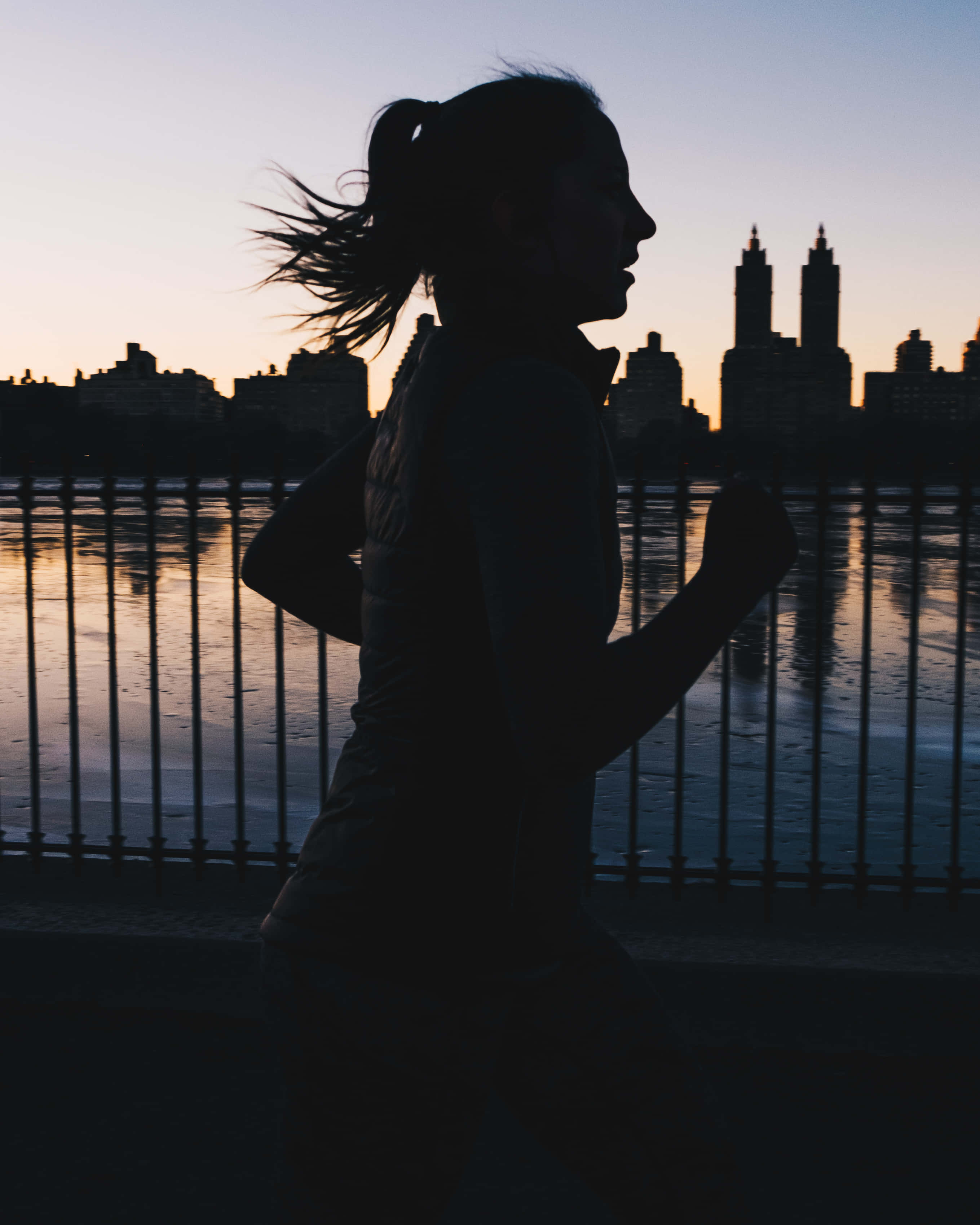 Run 桌布 壁紙 — 日落時分，一名女子沿著海濱欄杆慢跑的剪影，背景是城市天際線。
