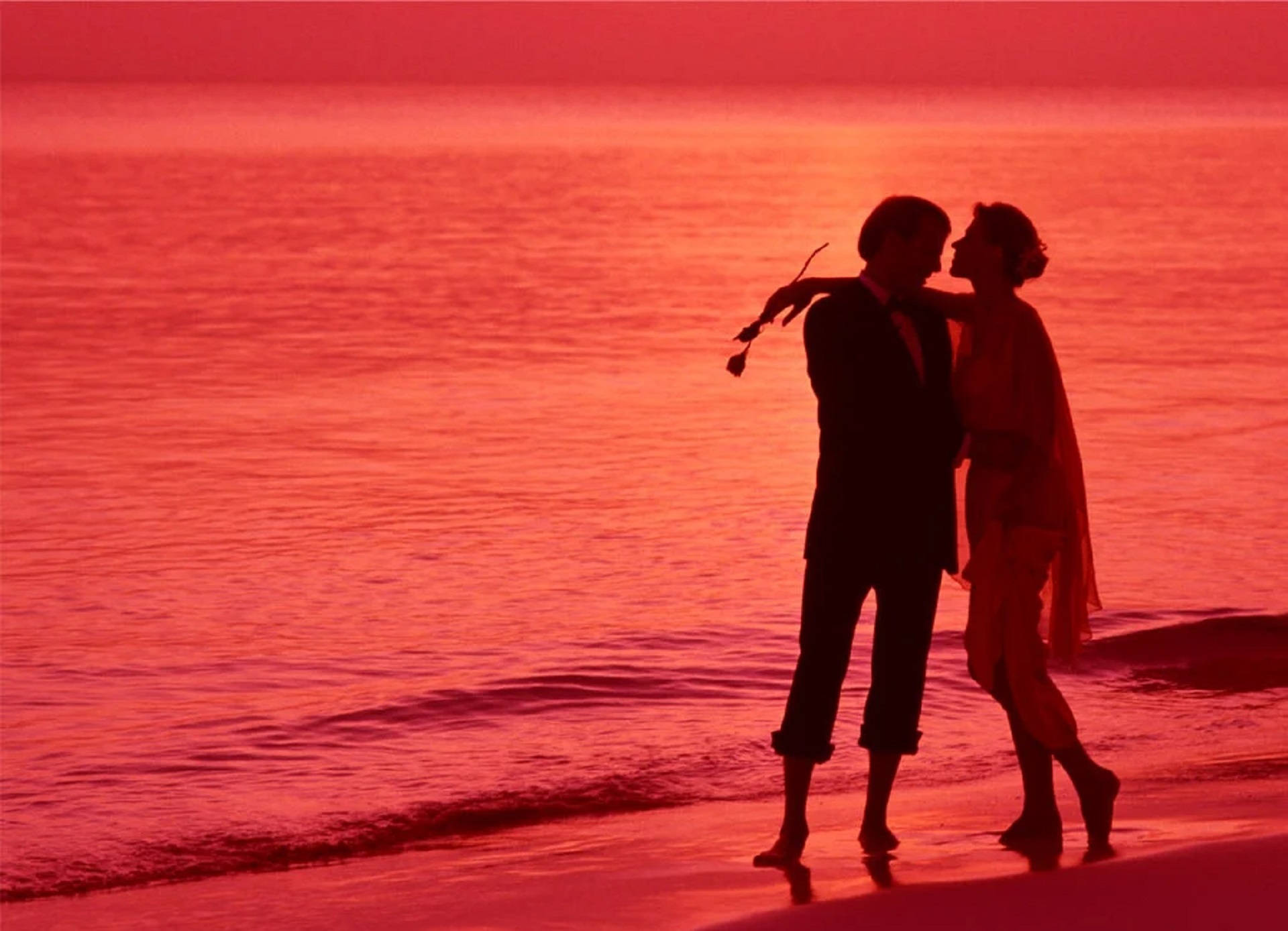 Romantic Love 桌布 壁紙 — 日落時分，一對剪影情侶在海灘上擁抱，海浪在深紅色調中閃耀。