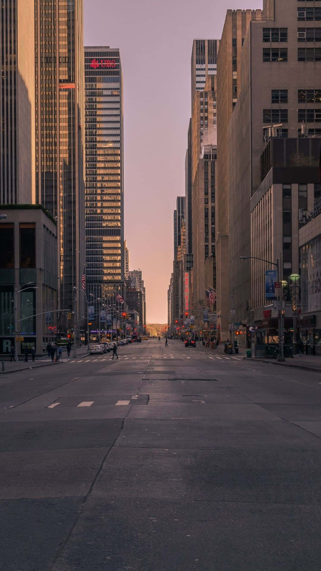 Android Ny City 桌布 壁紙 — 日落時分，一條寬闊的城市街道兩旁高聳的摩天大樓林立，溫暖的金色光芒投下長長的影子。