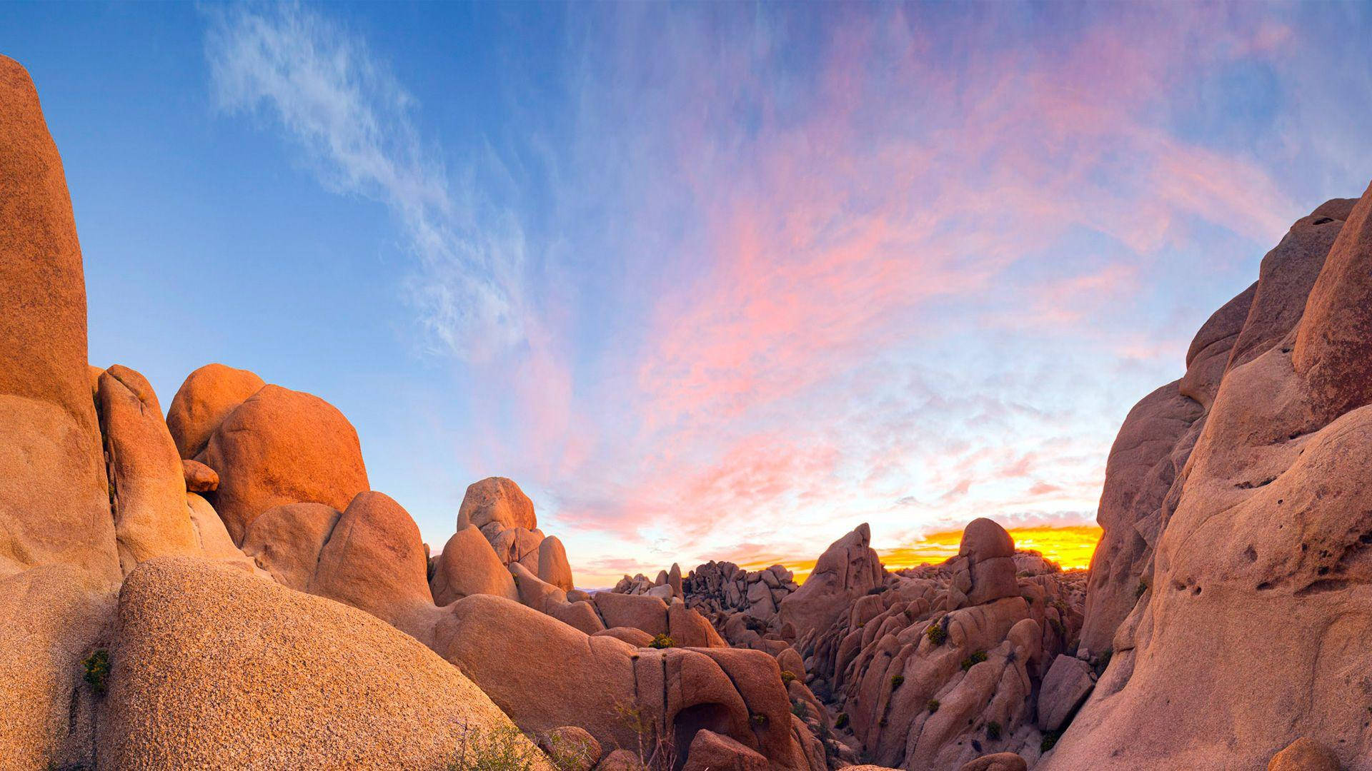 Joshua Tree National Park 桌布 壁紙 — 日落時分，在清澈的藍天下，粉紅色和金色的雲彩繪在一個由巨大圓形巨石組成的峽谷之上。