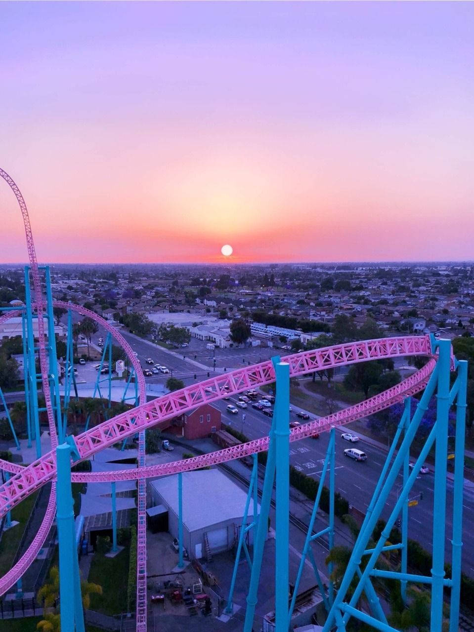 Roller Coaster 桌布 壁紙 — 日落時分，從空中俯瞰廣闊城市景觀上充滿活力的粉紅色和藍綠色雲霄飛車。