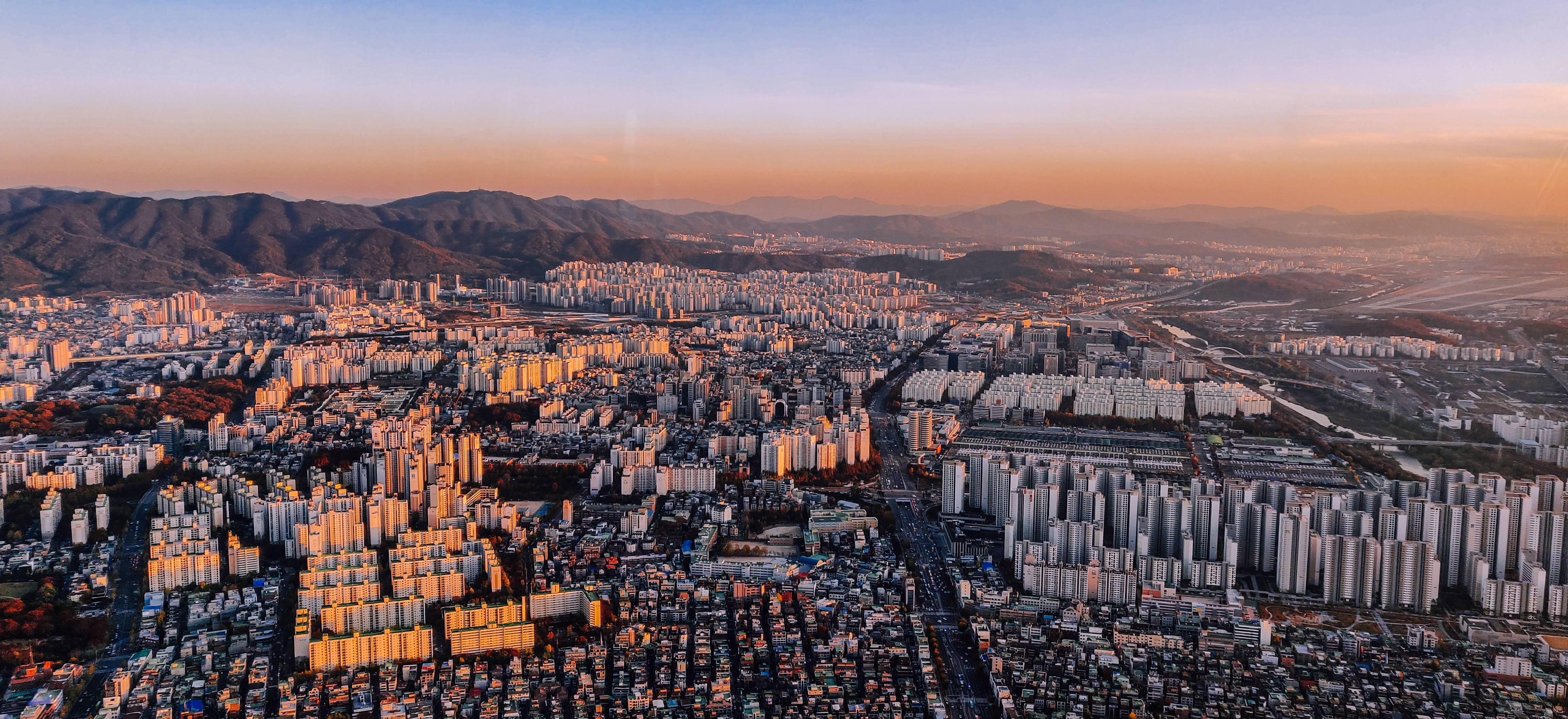首爾 桌布 壁紙 — 日落時分，從高空俯瞰一座廣闊的城市，金色陽光灑滿高樓大廈和遠山，天空呈現柔和的橙色。