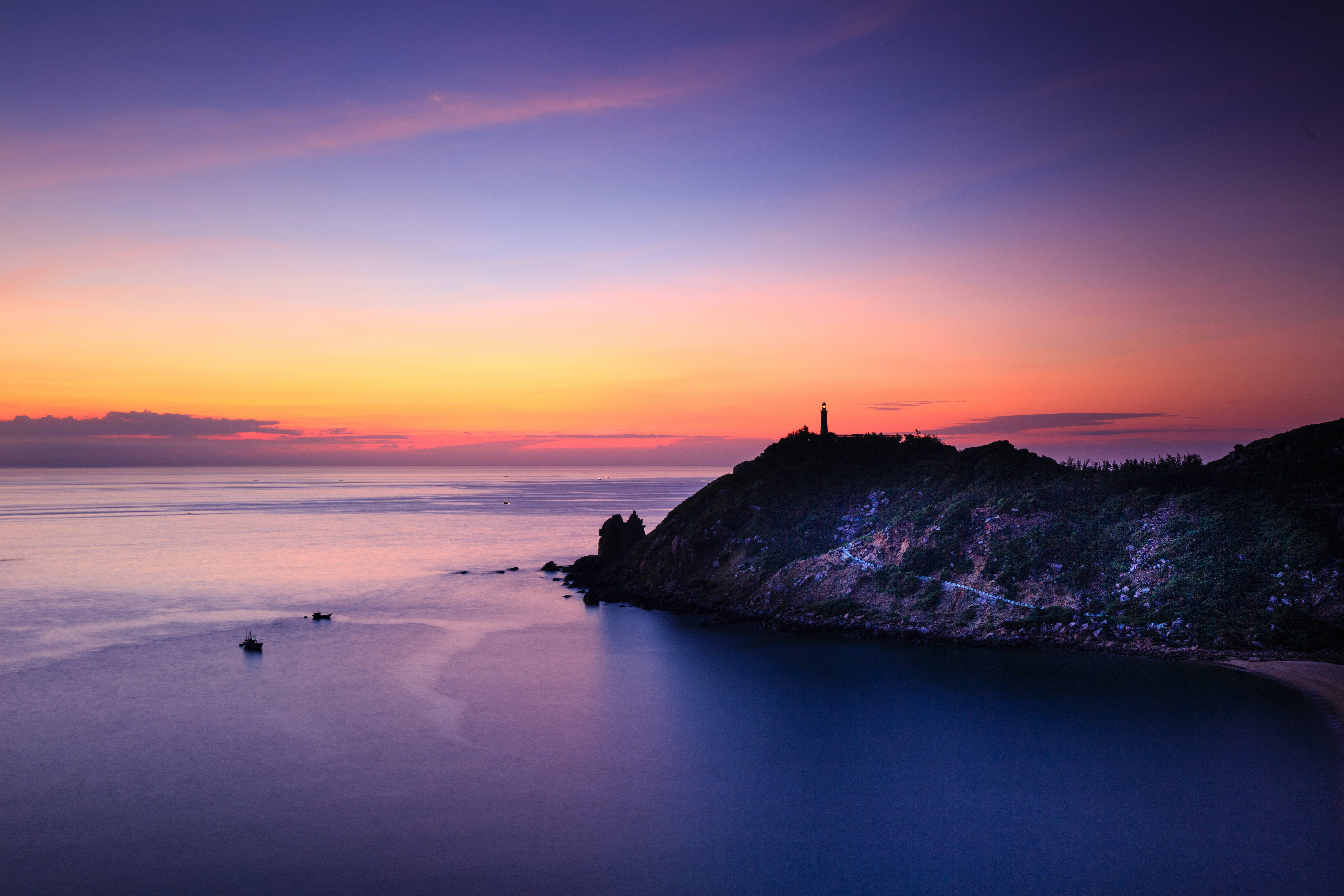 View 桌布 壁紙 — 日落時分，懸崖上的燈塔在平靜的海水和紫色的天空上投射出溫暖的光芒。