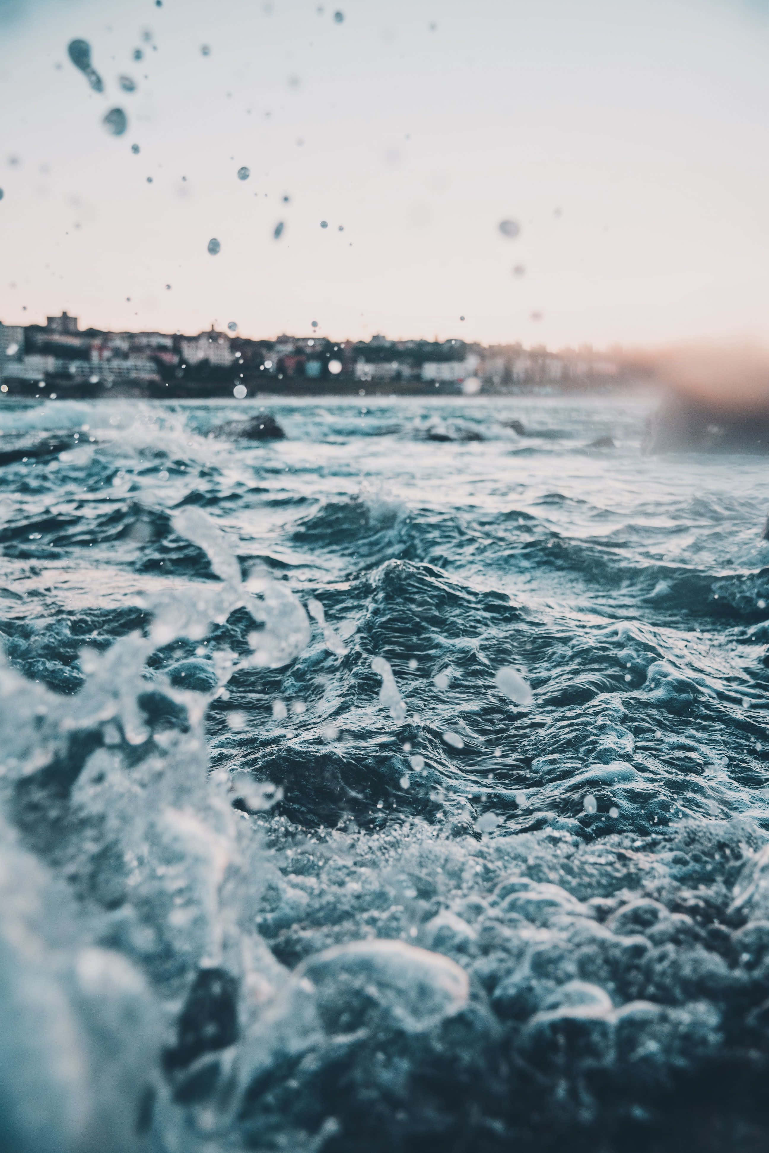 Ocean Aesthetic Phone 桌布 壁紙 — 日落時分，朦朧的海岸小鎮背景下，海浪拍打，水滴懸浮在空中的特寫。