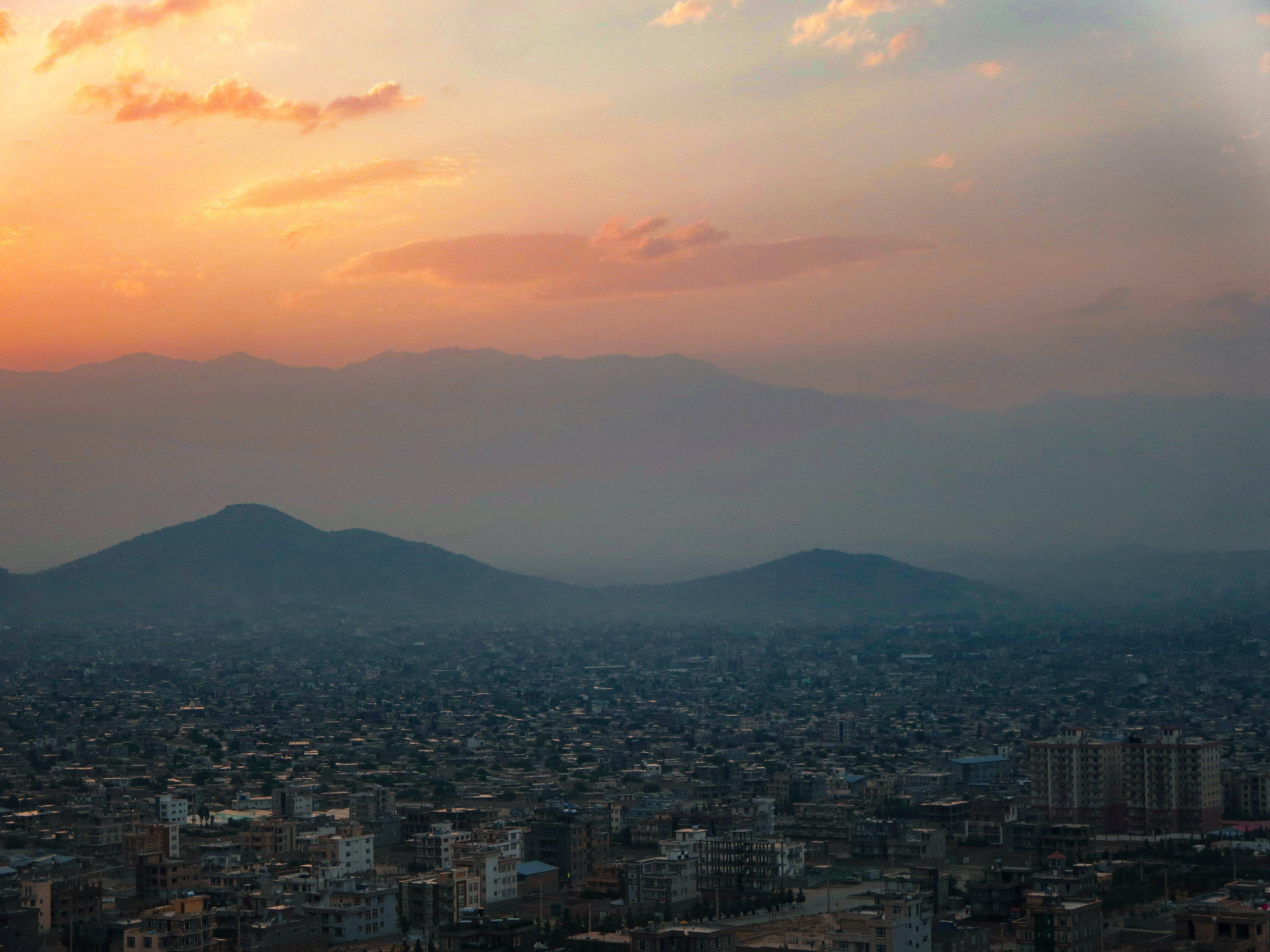 Kabul 桌布 壁紙 — 日落時分，橙色雲層覆蓋層層山脈，下方密集的城市建築，形成廣闊的城市景觀。