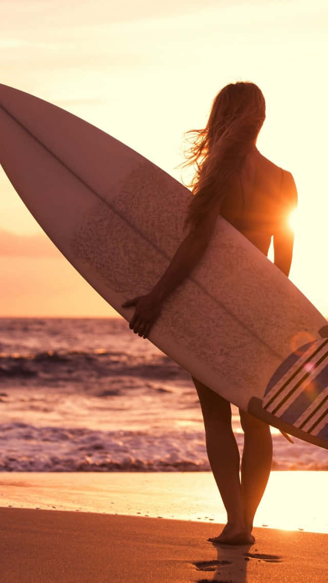 Surfing iPhone 桌布 壁紙 — 日落時分，沙灘上一位衝浪者手持衝浪板的剪影，背景是金色光芒與海浪。