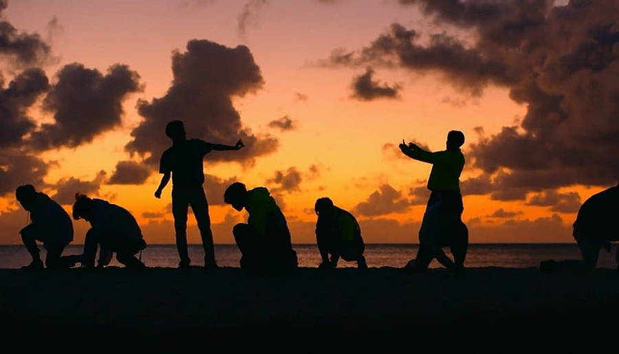 美學 桌布 壁紙 — 日落時分，沙灘上剪影人物擺出動態姿勢，背景是火橙色和紫色的雲彩。