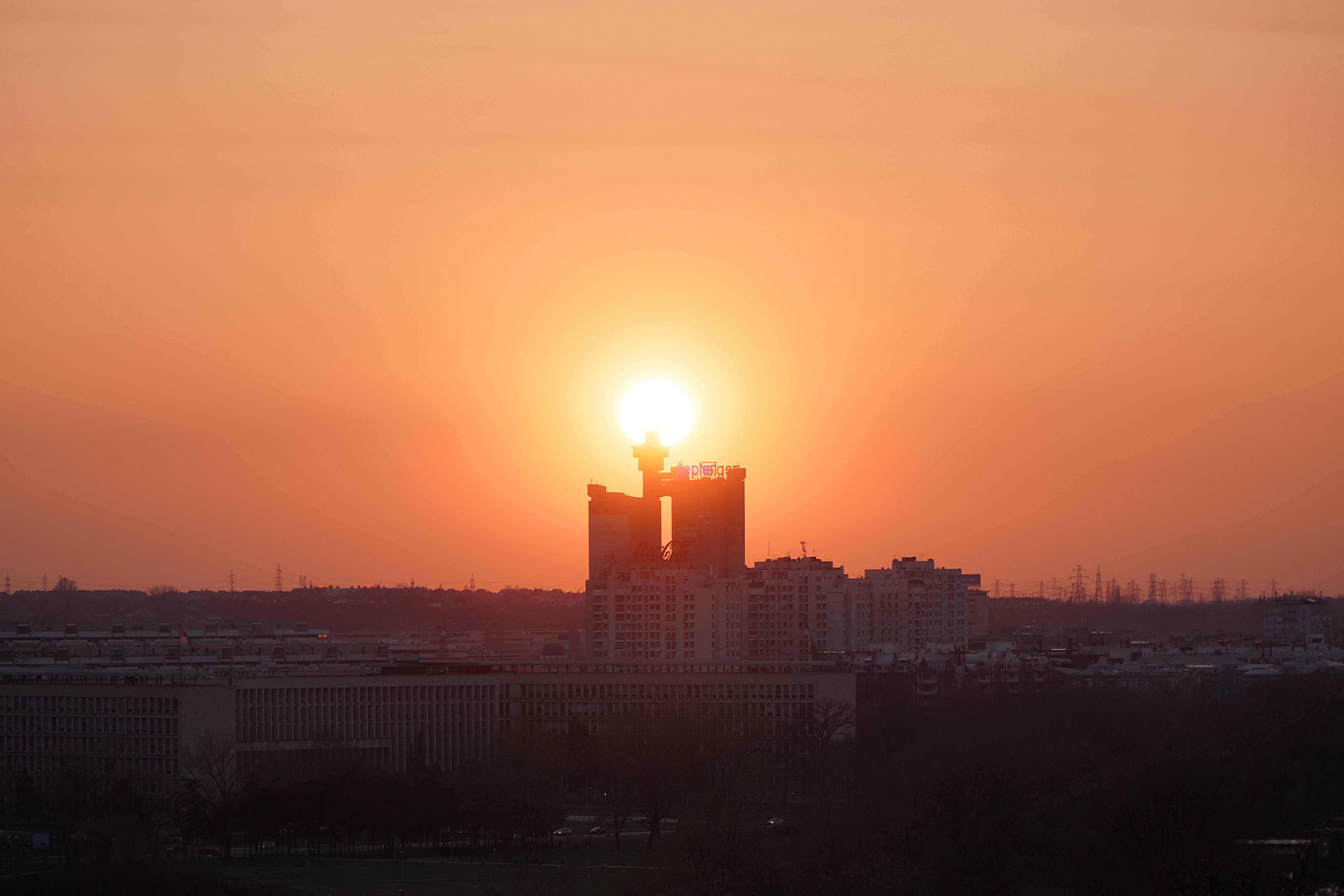 Serbia 桌布 壁紙 — 日落時分，現代城市天際線後方呈現橙色光芒，前景有剪影建築和光禿樹木。