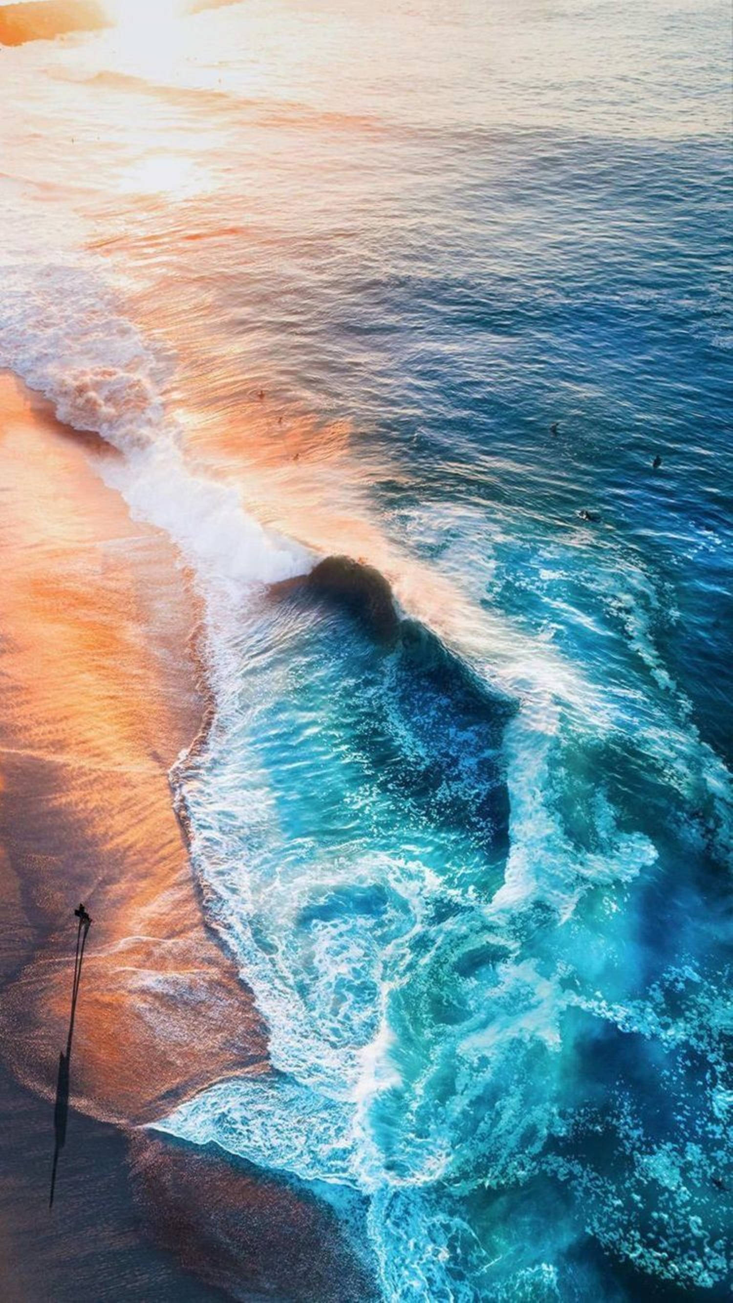 waves 桌布 壁紙 — 日落時分，翠綠海浪拍打金色沙灘的航拍視角，遠處有衝浪者。