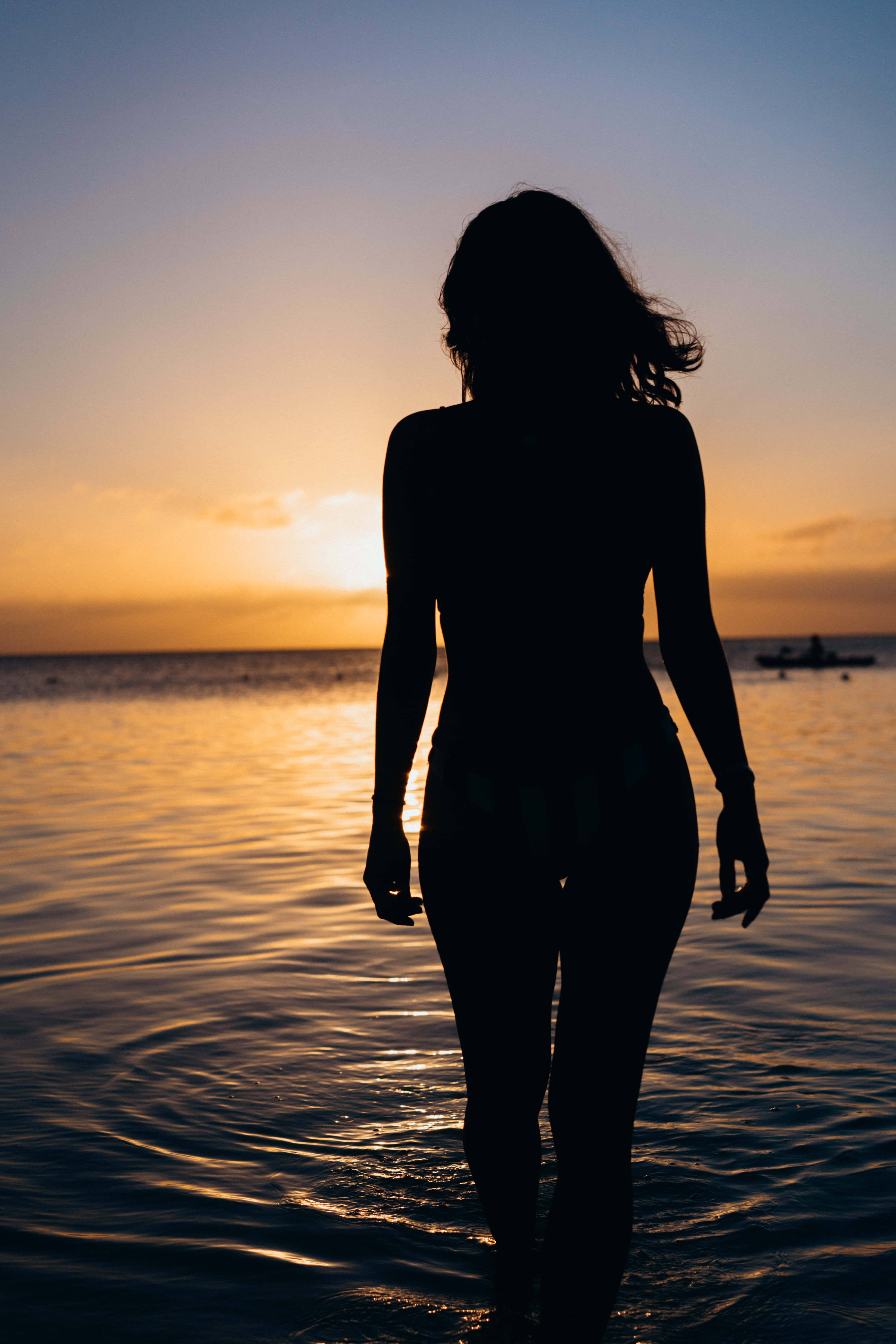 Silhouette 桌布 壁紙 — 日落時分，金色光芒映照海洋，一位女性赤腳在淺水中行走的剪影。