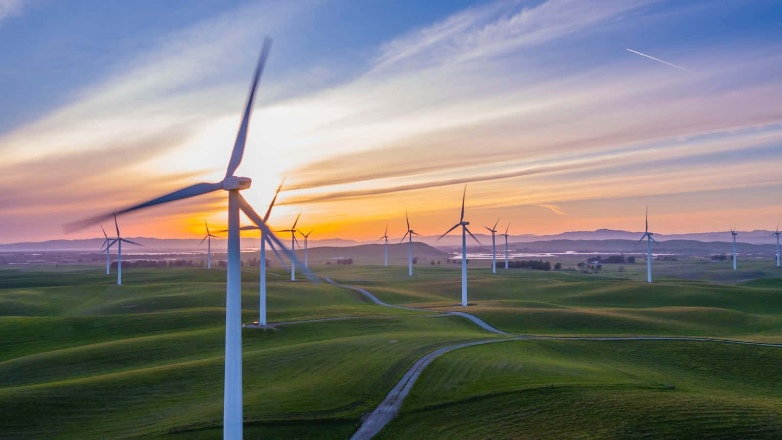 Renewable Energy 桌布 壁紙 — 日落時分，風力渦輪機在連綿起伏的綠色山丘上旋轉的鳥瞰圖，帶有條紋雲和遠處的山脈。