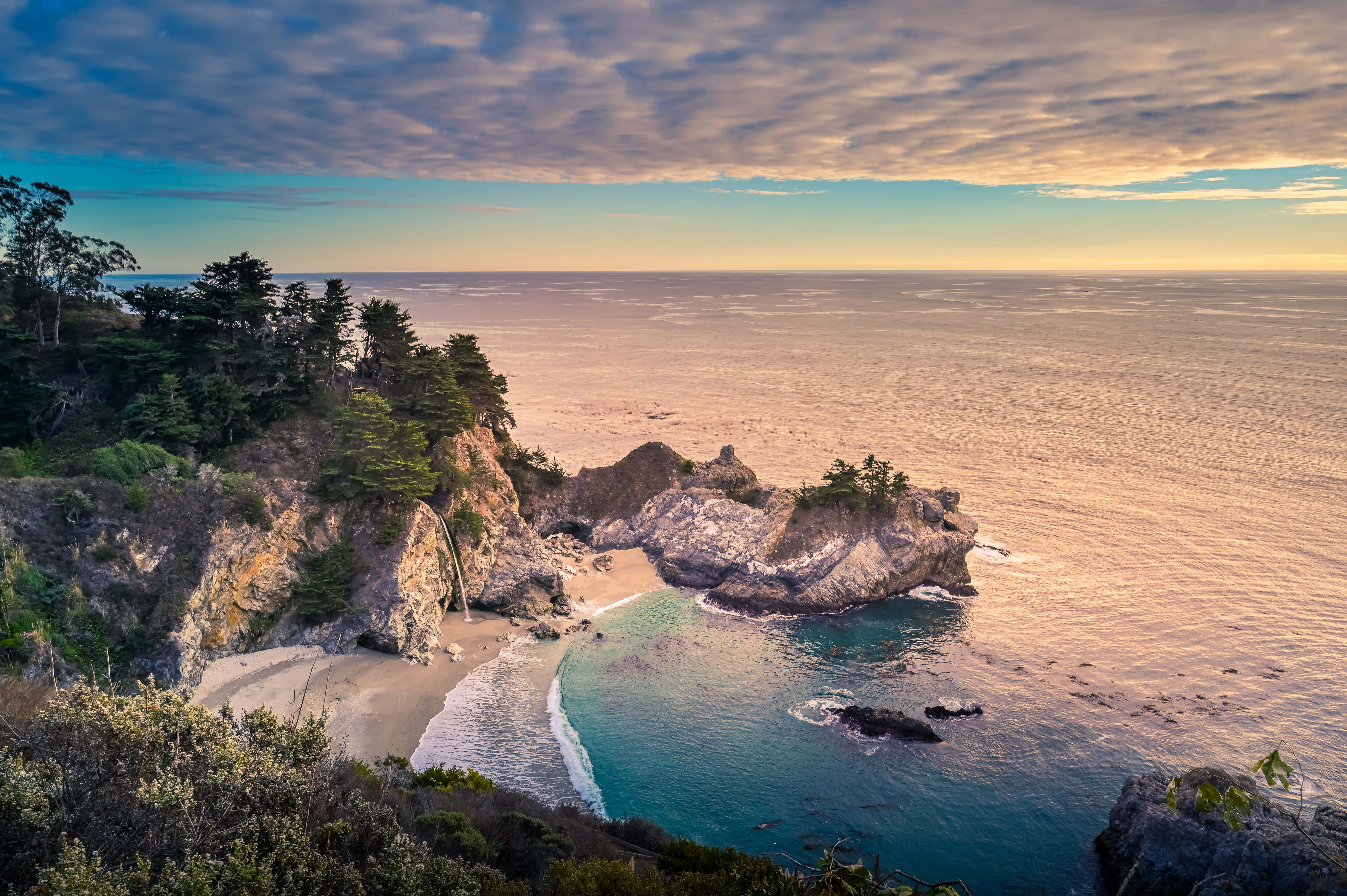 Big Sur 桌布 壁紙 — 日落時分一個隱蔽的小海灣，碧綠的海水輕拍著沙灘，四周環繞著崎嶇的懸崖和常綠樹木。