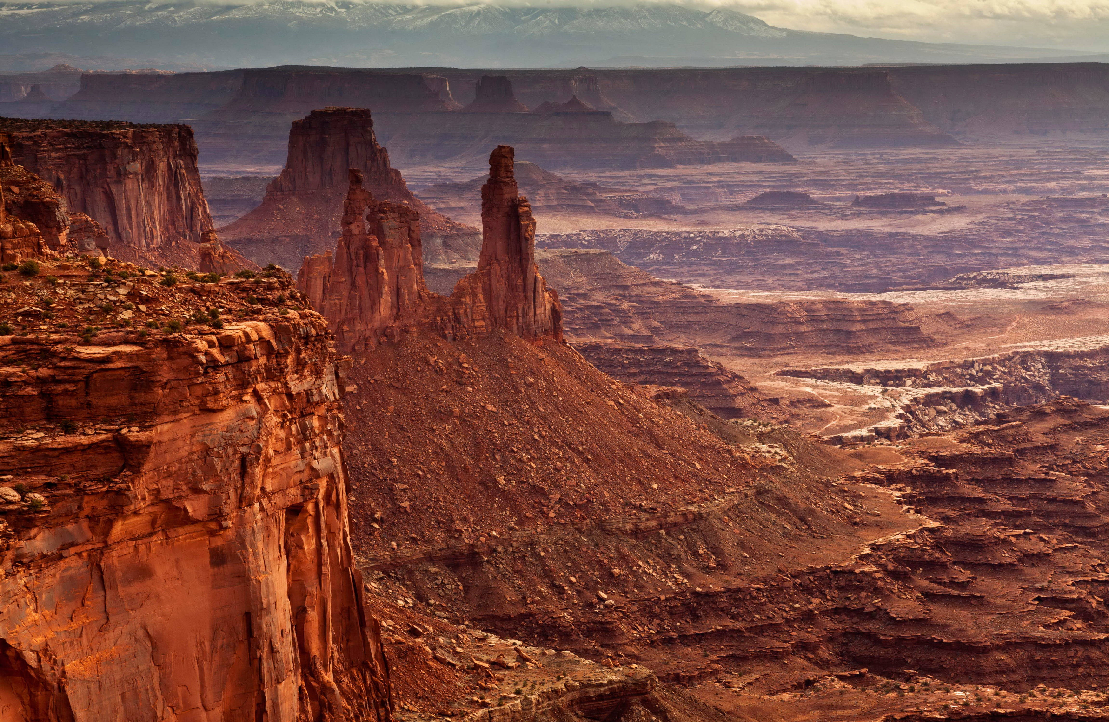 Canyonlands National Park 桌布 壁紙 — 日落時分令人驚嘆的沙漠峽谷，高聳的紅色岩層和遠處白雪覆蓋的山脈在柔和的金色天空下。