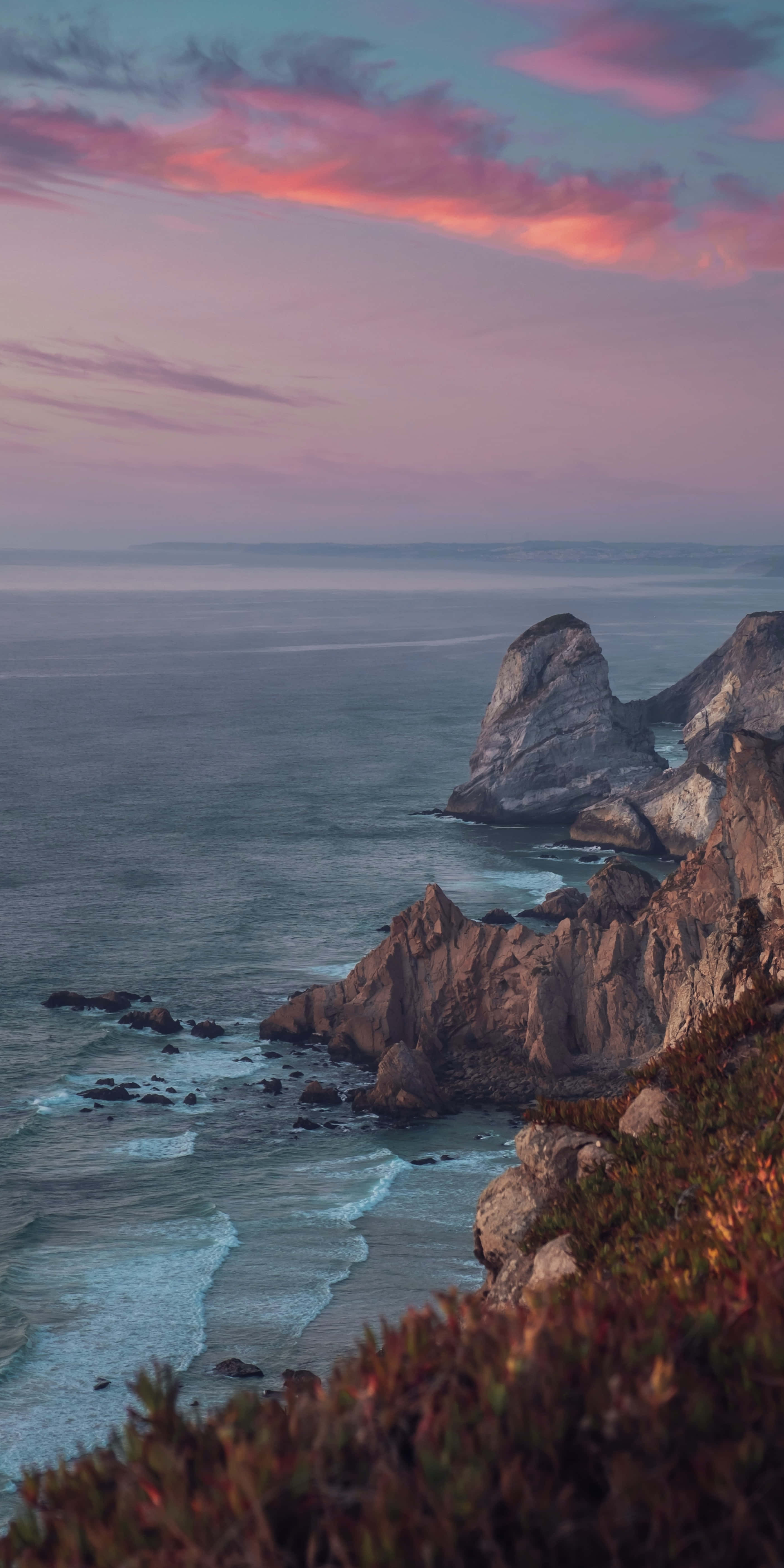 Calm Aesthetic 桌布 壁紙 — 日落時分令人驚嘆的海岸懸崖，崎嶇岩石上方有粉紅色和紫色的雲彩，下方有拍打的波浪。