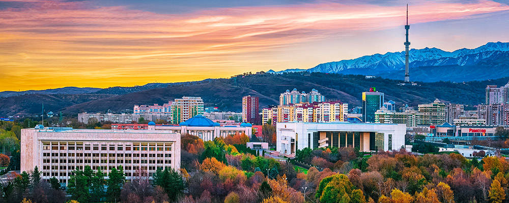 Kazakhstan 桌布 壁紙 — 日落時分充滿活力的城市景觀，前景有秋樹，現代建築和山脈在火熱的橙色和粉紅色天空下。