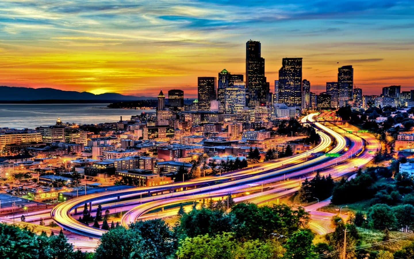 Washington 桌布 壁紙 — 日落時分充滿活力的城市景觀，發光的摩天大樓和蜿蜒穿過茂密綠化的車流光跡。