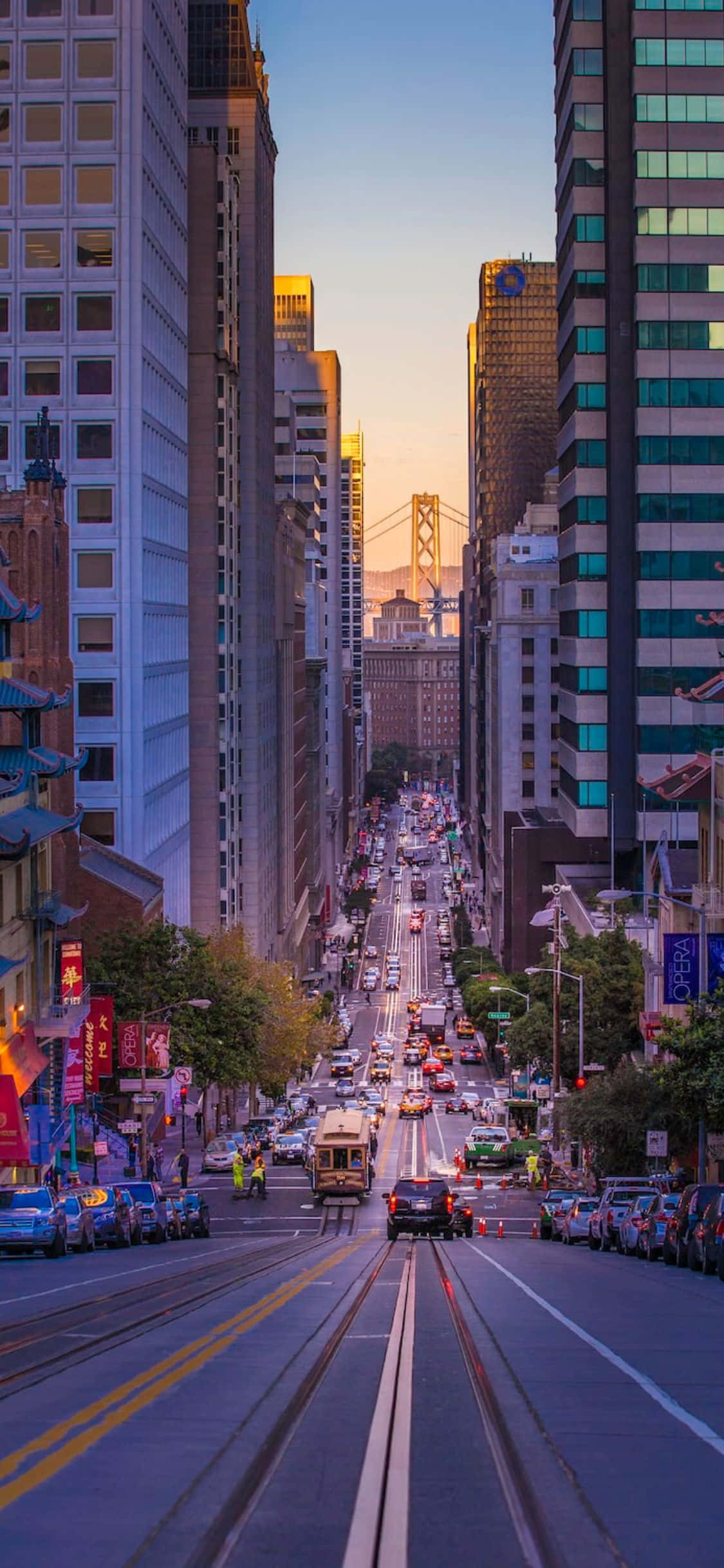 iPhone Xs California 桌布 壁紙 — 日落時分充滿活力的城市街道，高聳的摩天大樓框住了遠處金門大橋的景色。