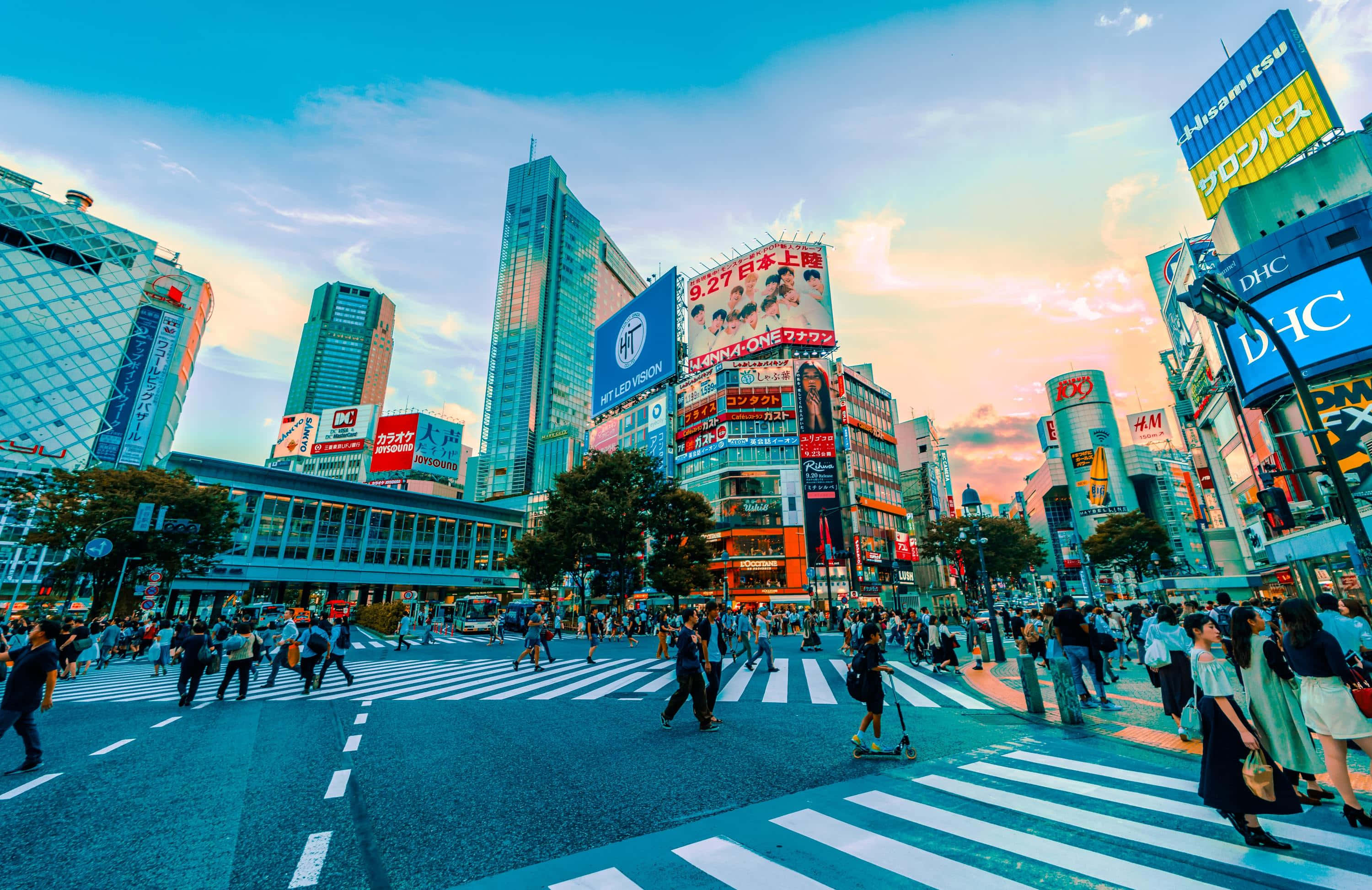東京 桌布 壁紙 — 日落時分充滿活力的東京街道，人群在閃爍的霓虹燈招牌和高聳的摩天大樓下穿越斑馬線。