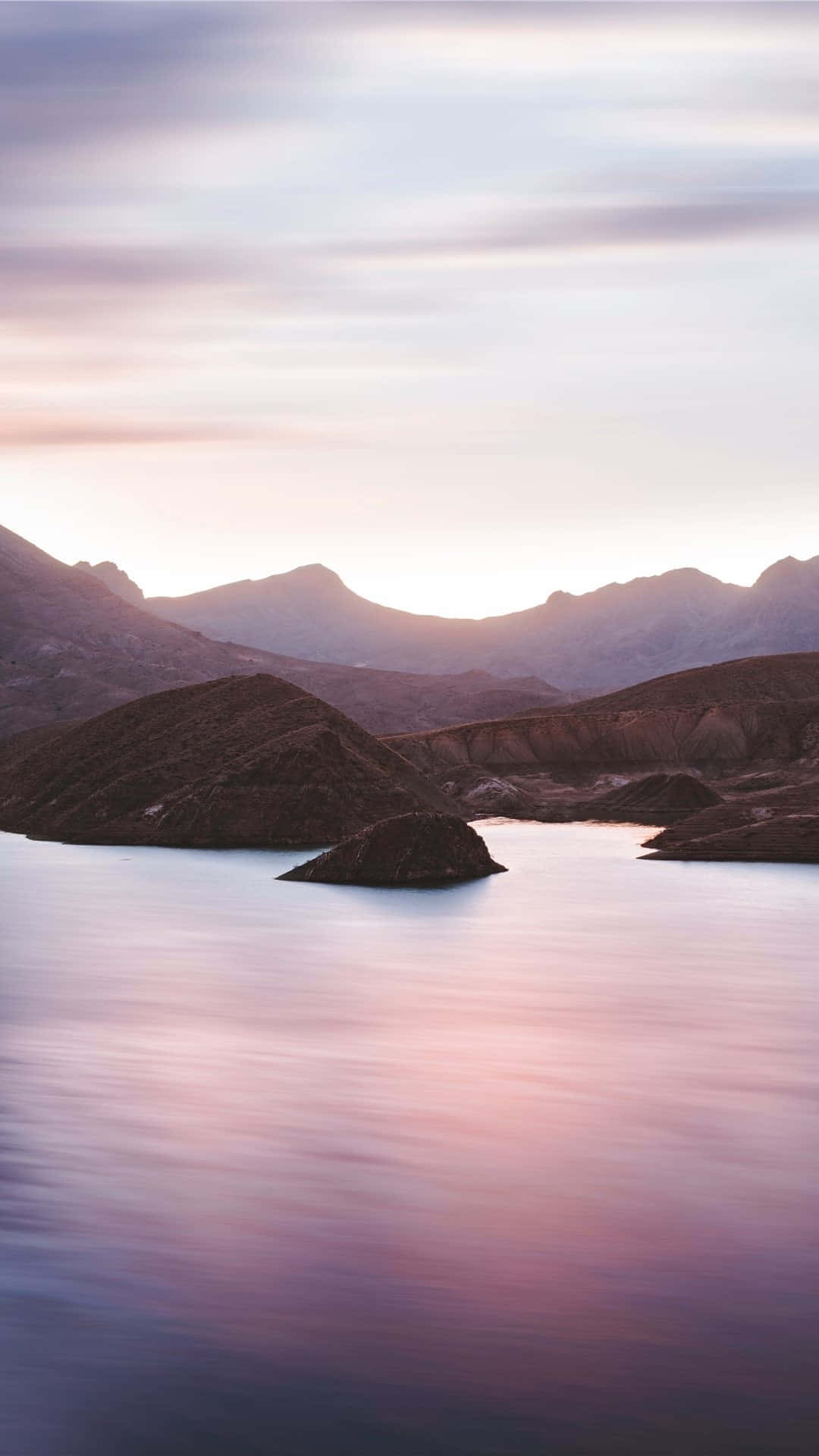 Calming iPhone 桌布 壁紙 — 日落時分寧靜的山間湖泊，柔和的粉紅和紫色色調映照在平靜的水面上，四周環繞著崎嶇的山丘。
