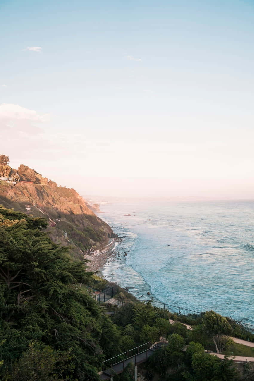 Beach Vacation 桌布 壁紙 — 日落時分寧靜的海岸懸崖，青綠色的海浪拍打岩石，下方是茂密的綠色植物。
