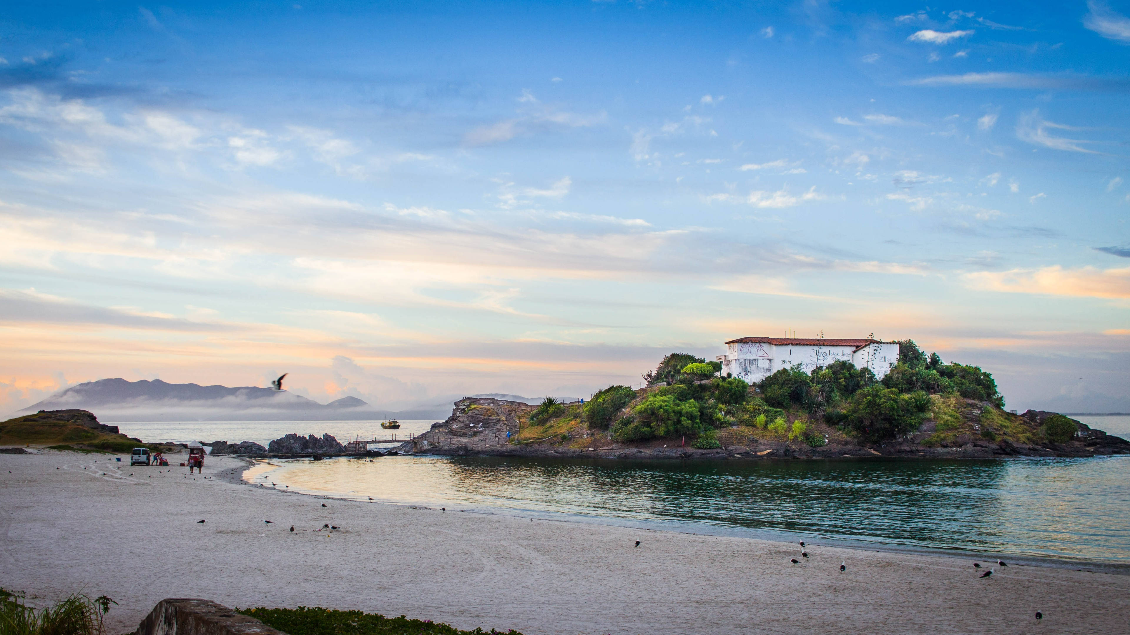 rio 桌布 壁紙 — 日落時分寧靜的海岸景色，白色小教堂坐落於岩石島上，四周環繞平靜水域和沙灘。