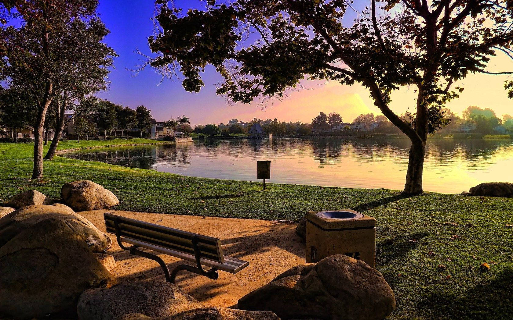 Bench 桌布 壁紙 — 日落時分寧靜的湖邊公園，木製長椅隱藏在岩石和樹木之間，平靜的水面反射著金色的光芒。