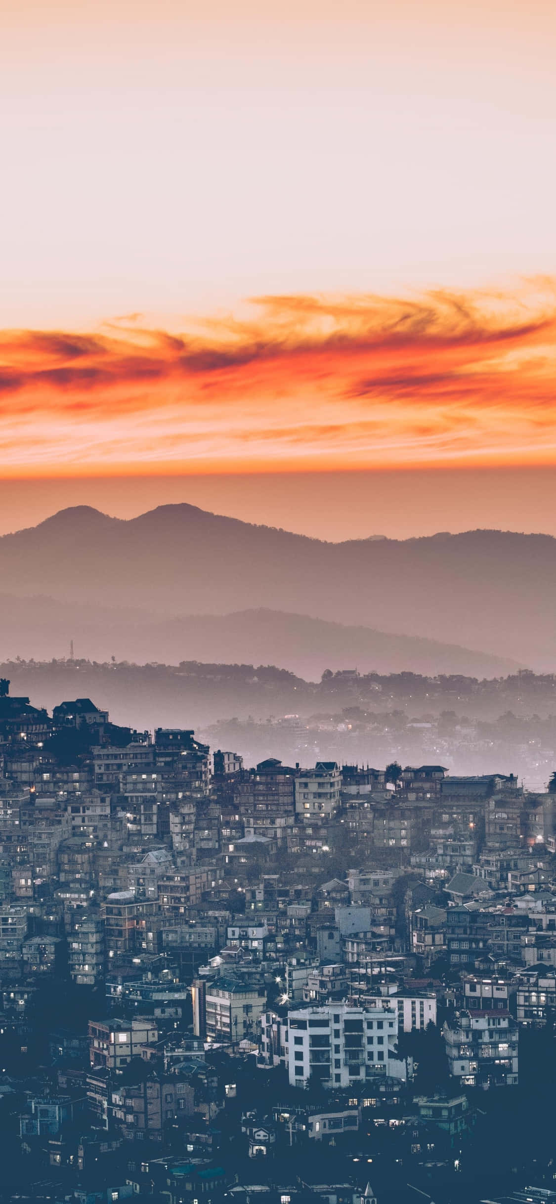 City Sunset iPhone 桌布 壁紙 — 日落時分廣闊的城市景觀，橙色雲層籠罩著霧氣繚繞的山巒，下方窗戶閃爍著光芒。