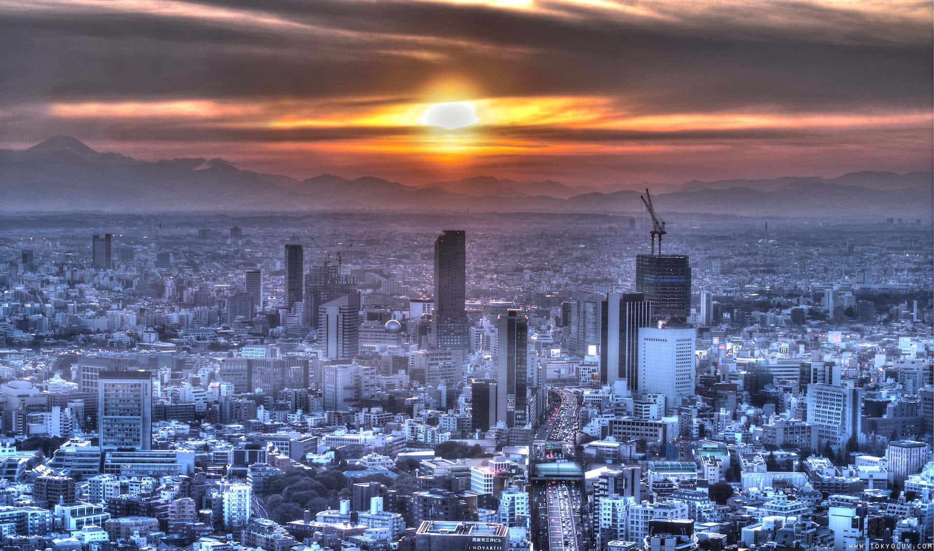 東京 桌布 壁紙 — 日落時分廣闊的城市景觀，橙色雲彩籠罩在藍色調的摩天大樓和遠山之上。