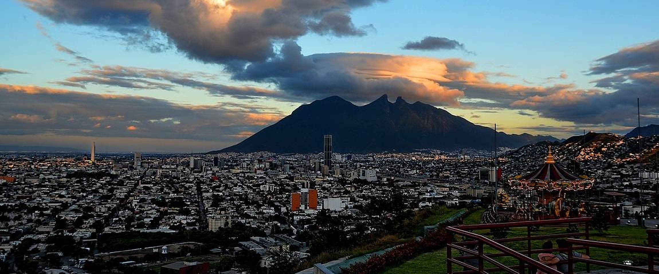 Monterrey 桌布 壁紙 — 日落時分廣闊的城市景觀，背後聳立著雄偉的山脈，上方雲彩閃耀著橙色和粉紅色。