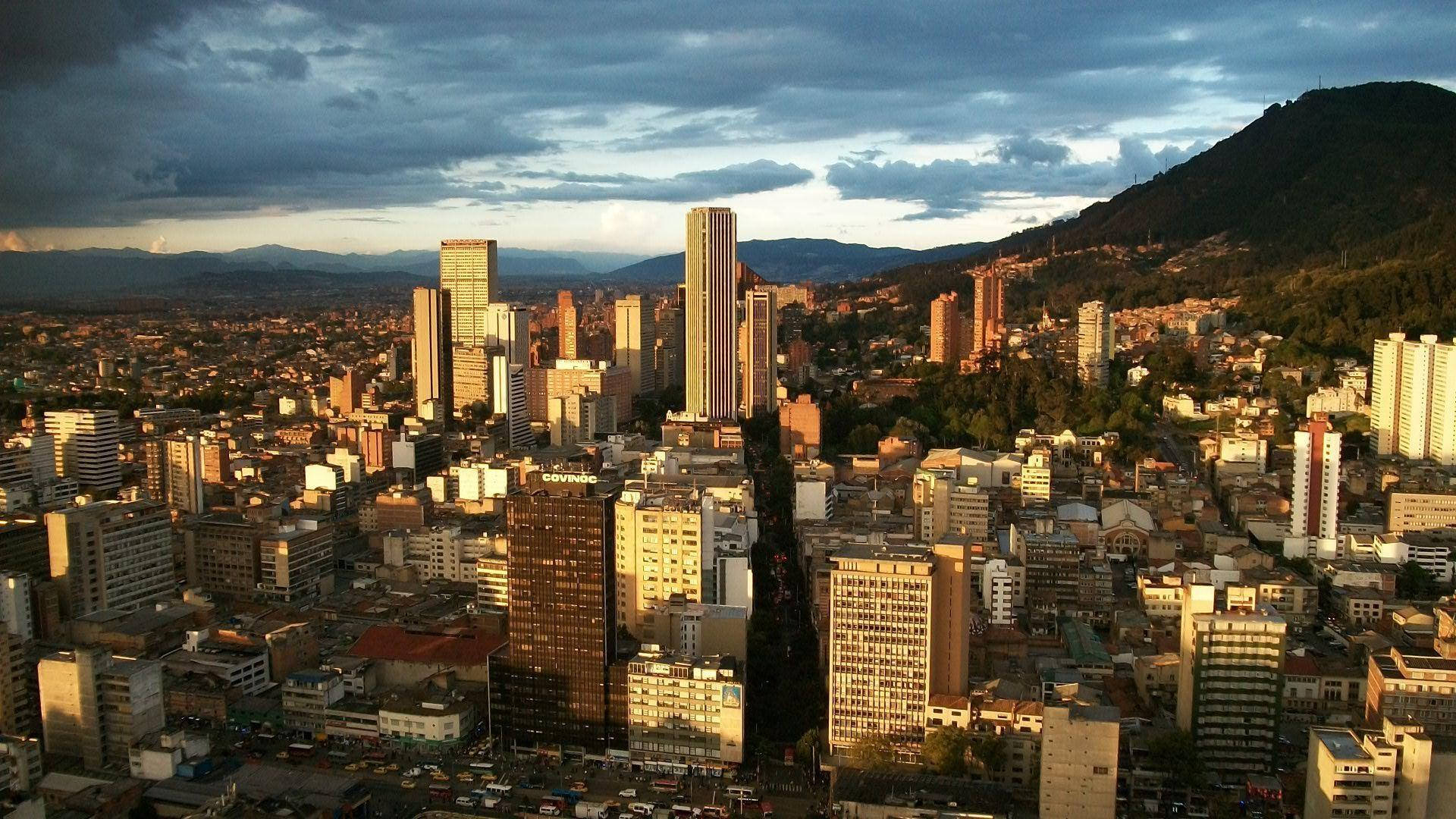 Bogota 桌布 壁紙 — 日落時分廣闊的城市景觀，金色摩天大樓和密集的住宅建築依偎在森林覆蓋的山脈下，天空中有戲劇性的雲彩。