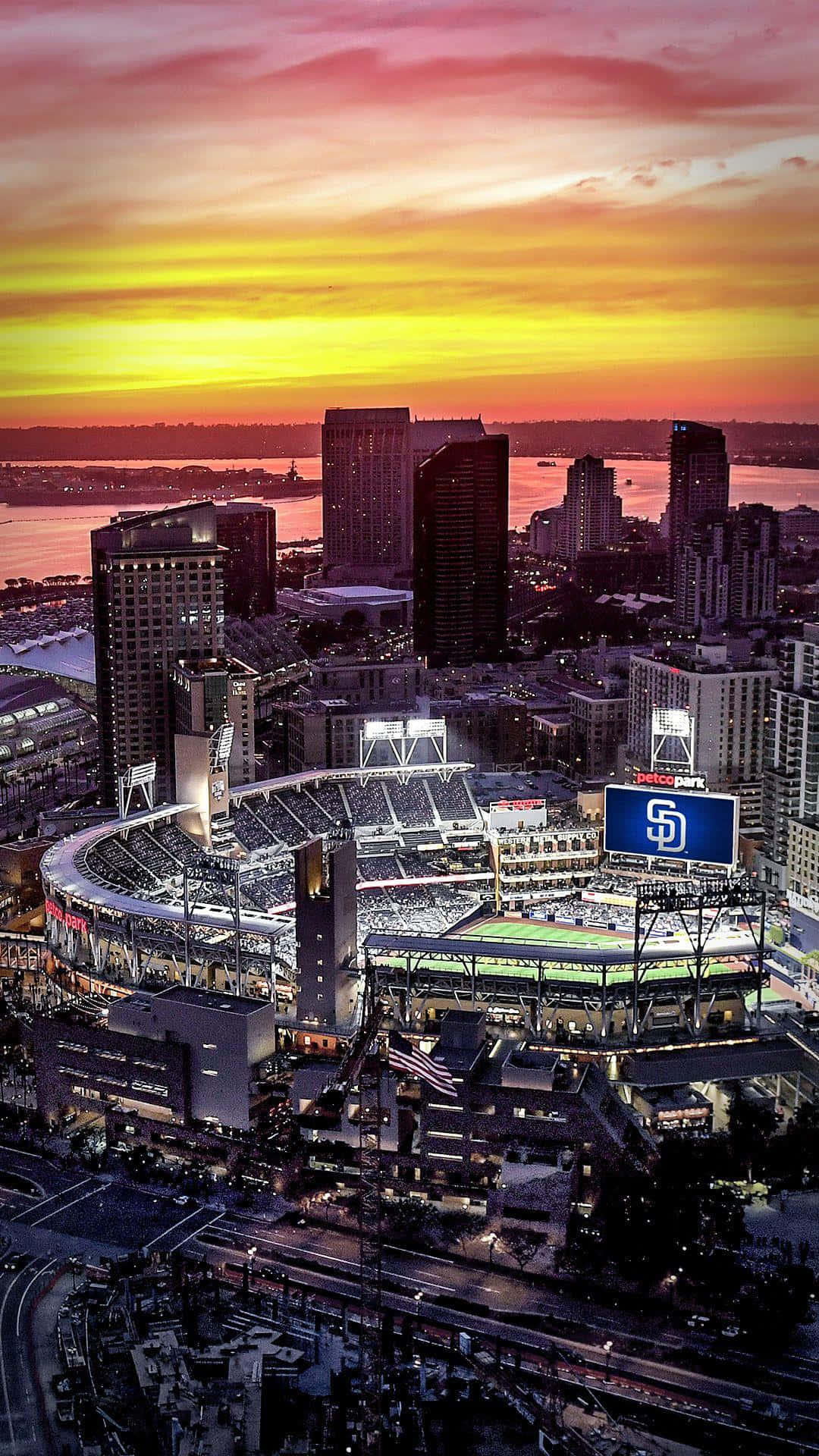 Stadium 桌布 壁紙 — 日落時分棒球場的鳥瞰圖，城市景觀和水面上方有火熱的橙色和粉紅色天空。