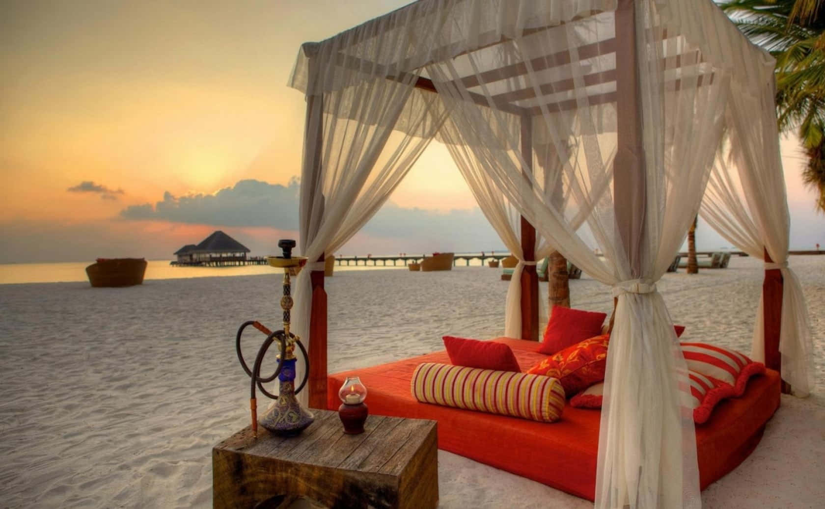 Beach Daybed 桌布 壁紙 — 日落時分沙灘上白色天篷下的紅色躺椅，木桌上放著水煙壺。