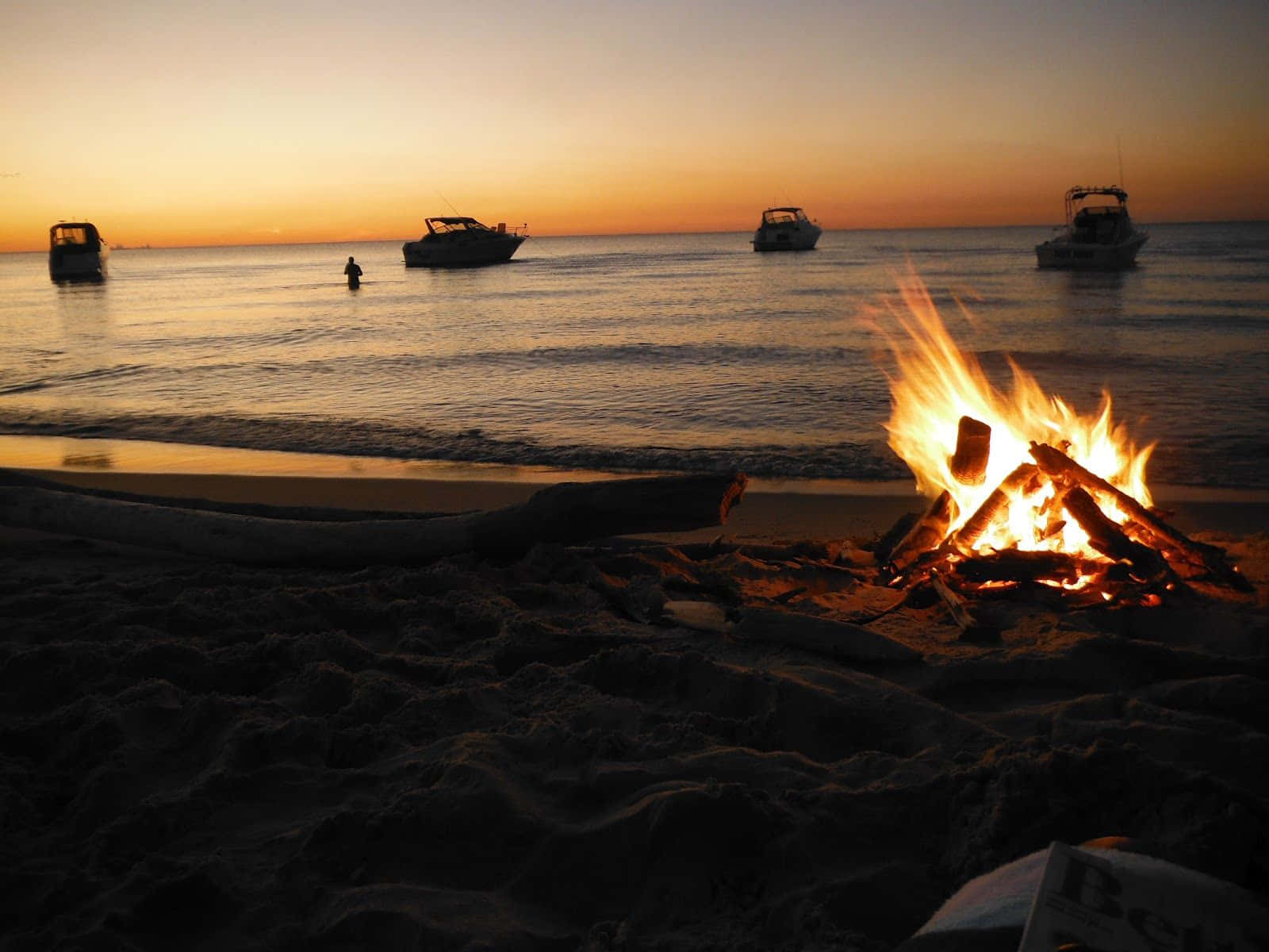 Beach Bonfire 桌布 壁紙 — 日落時分海灘上熊熊燃燒的營火，剪影般的船隻和海中孤獨的游泳者。