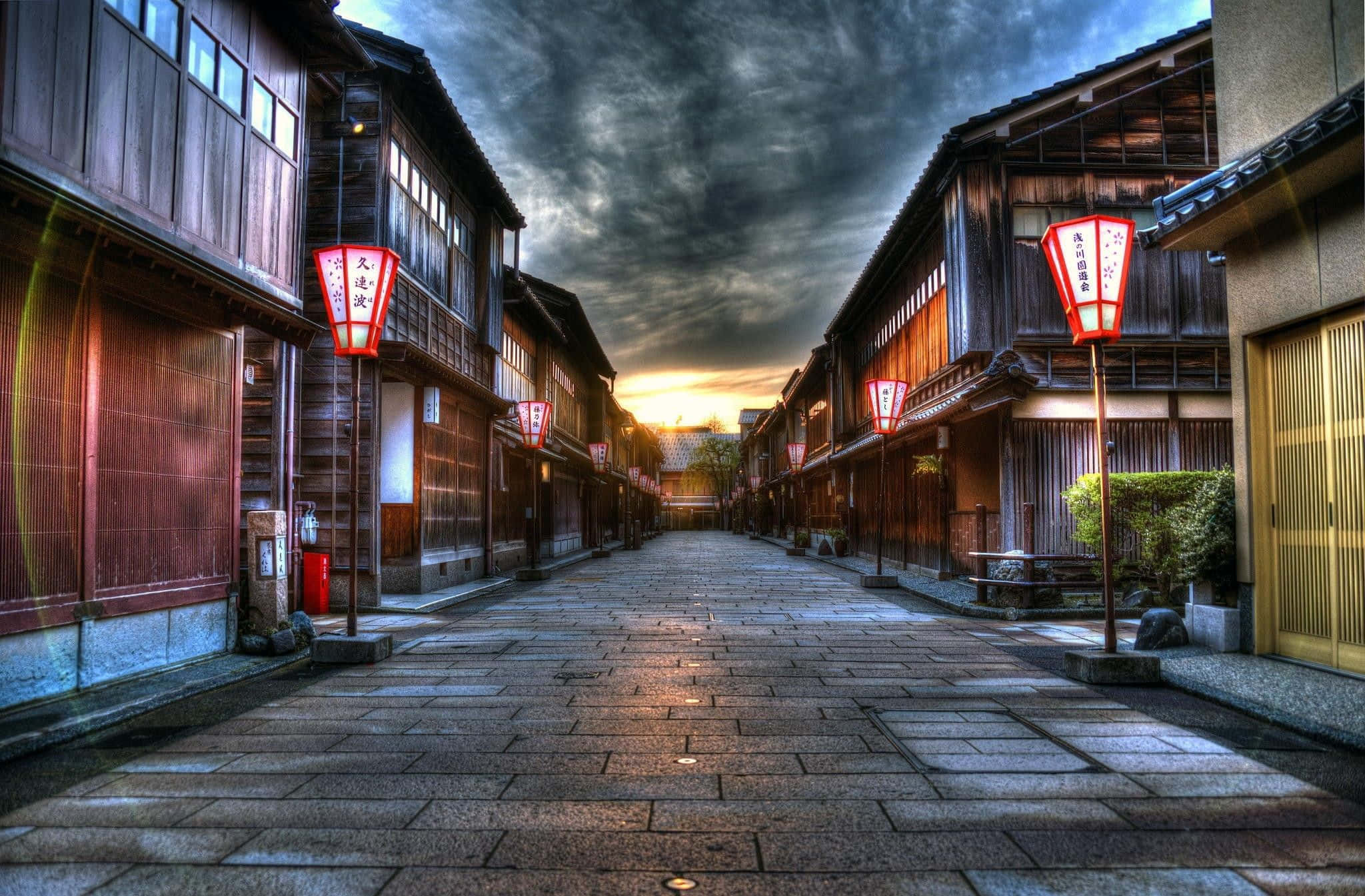 Road 桌布 壁紙 — 日落時分狹窄的傳統日本街道，紅色燈籠閃爍，木建築在戲劇性的多雲天空下。