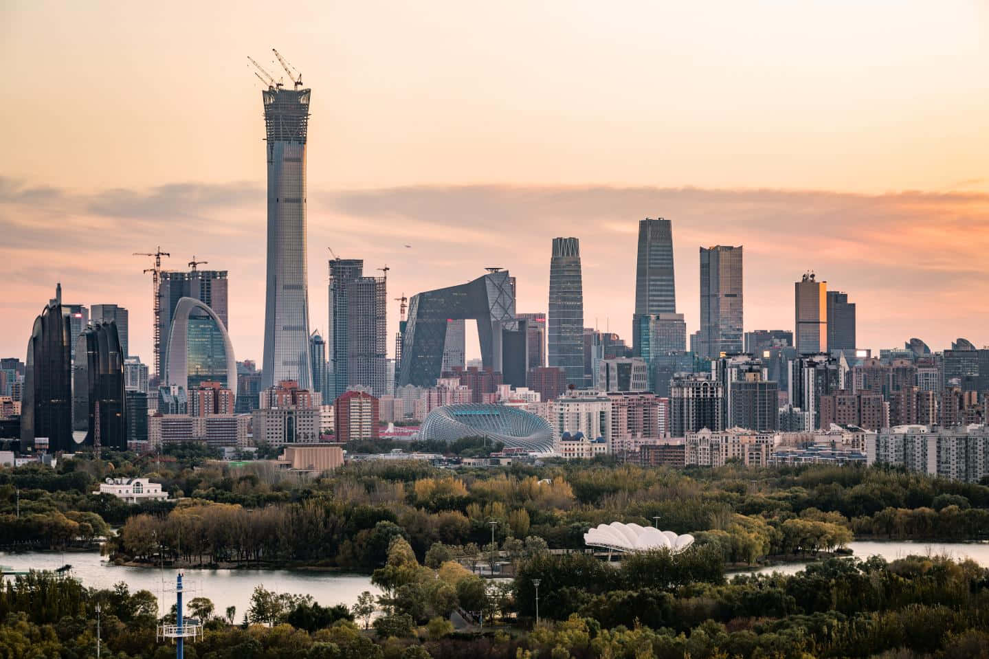 中國 桌布 壁紙 — 日落時分現代城市天際線的全景視圖，高樓大廈和建築起重機聳立在河流和綠色公園之上。