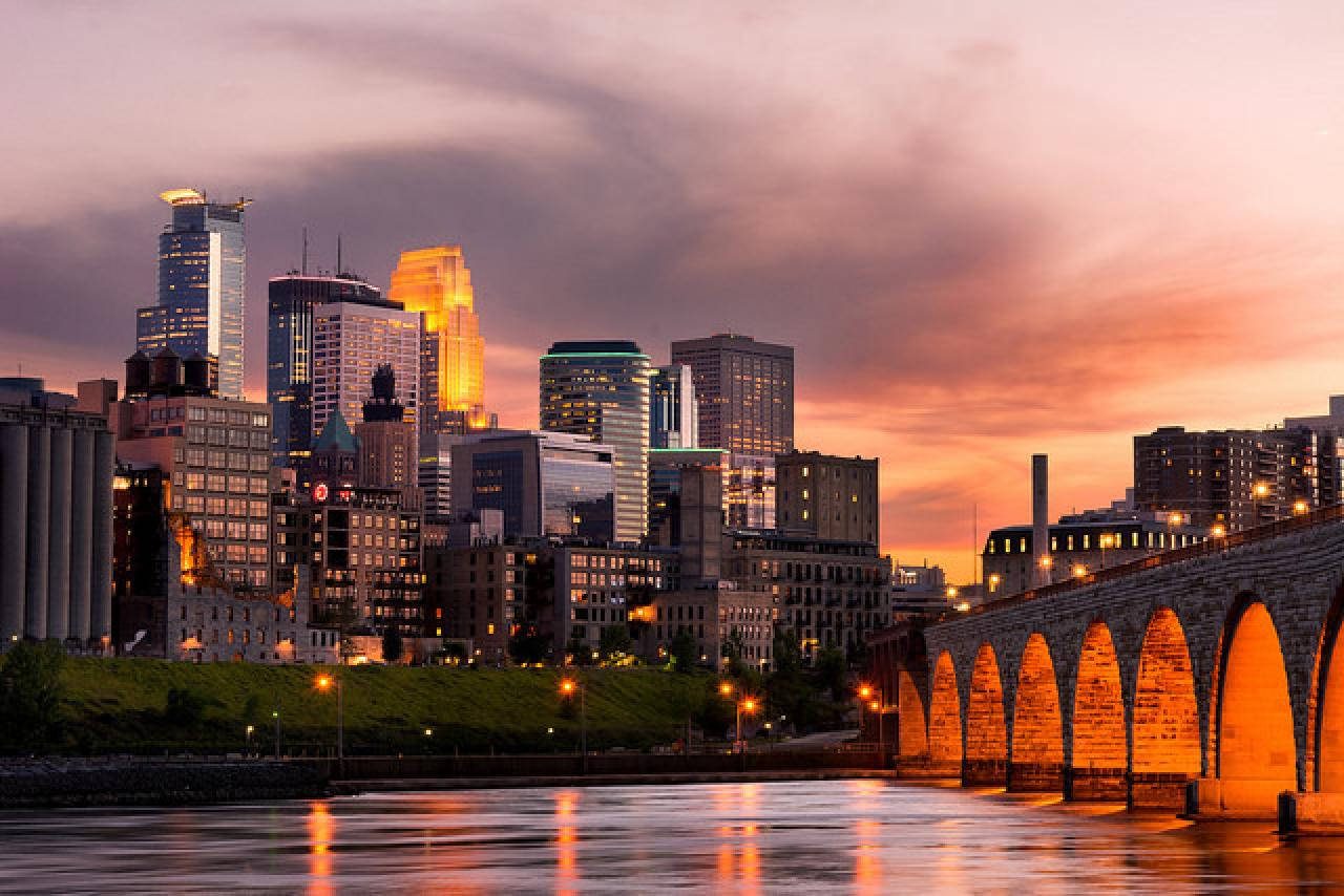 Minneapolis 桌布 壁紙 — 日落時分的城市天際線，一座石橋拱跨在水面上，倒映著溫暖的橙粉天色。