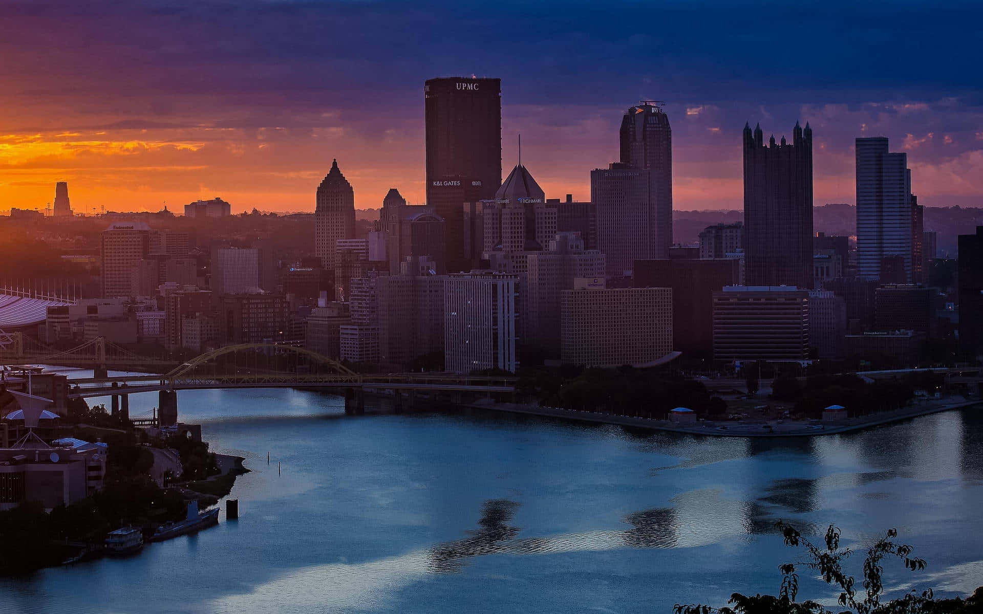Pittsburgh Skyline 桌布 壁紙 — 日落時分的城市天際線，橙色和紫色的天空倒映在下方平靜的河流上。