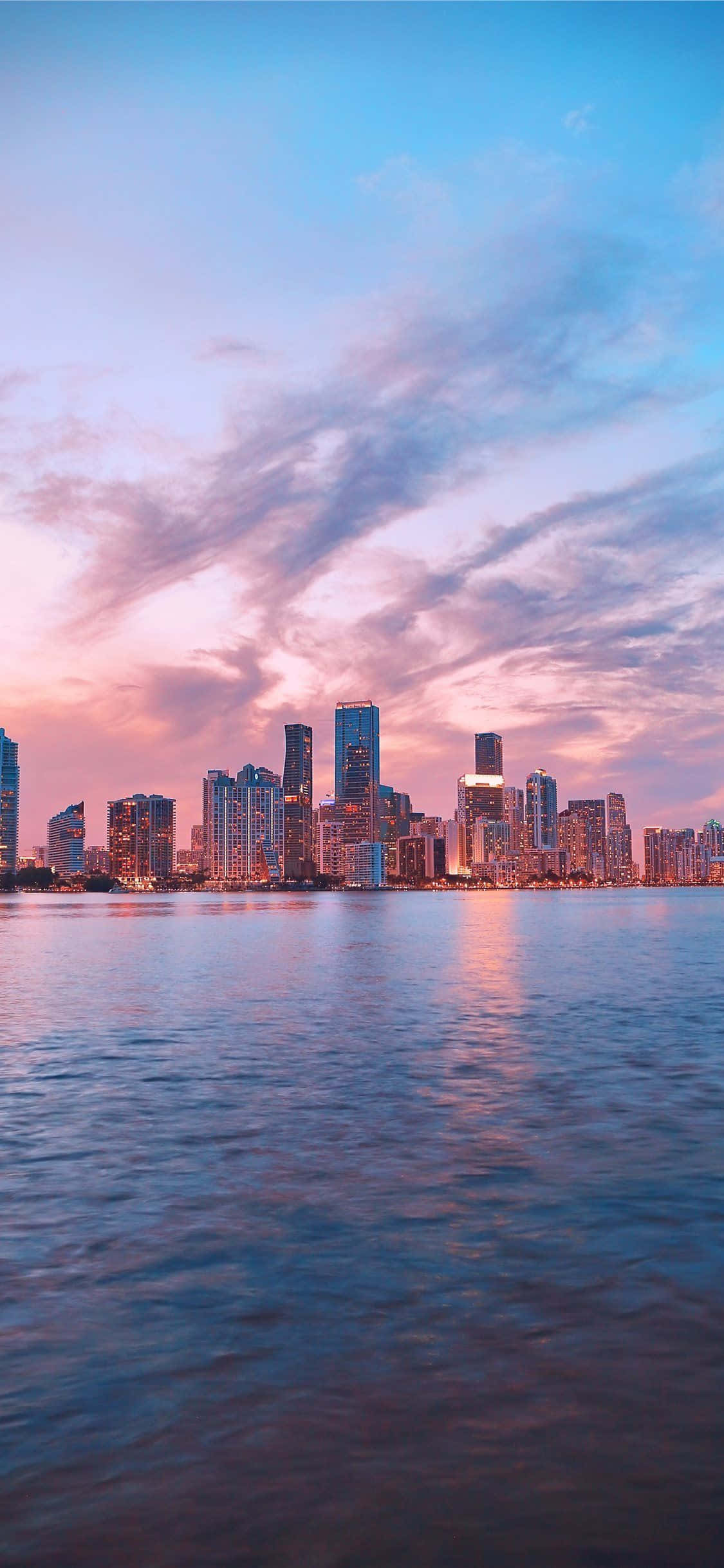 Miami iPhone 桌布 壁紙 — 日落時分的城市天際線，粉紅色和藍色的雲彩倒映在平靜的水面上。