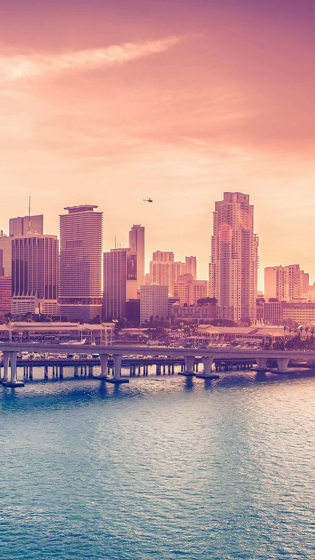 City Sunset iPhone 桌布 壁紙 — 日落時分的城市天際線，高樓大廈在粉紅色和橙色的天空下倒映在平靜的藍色水面上。