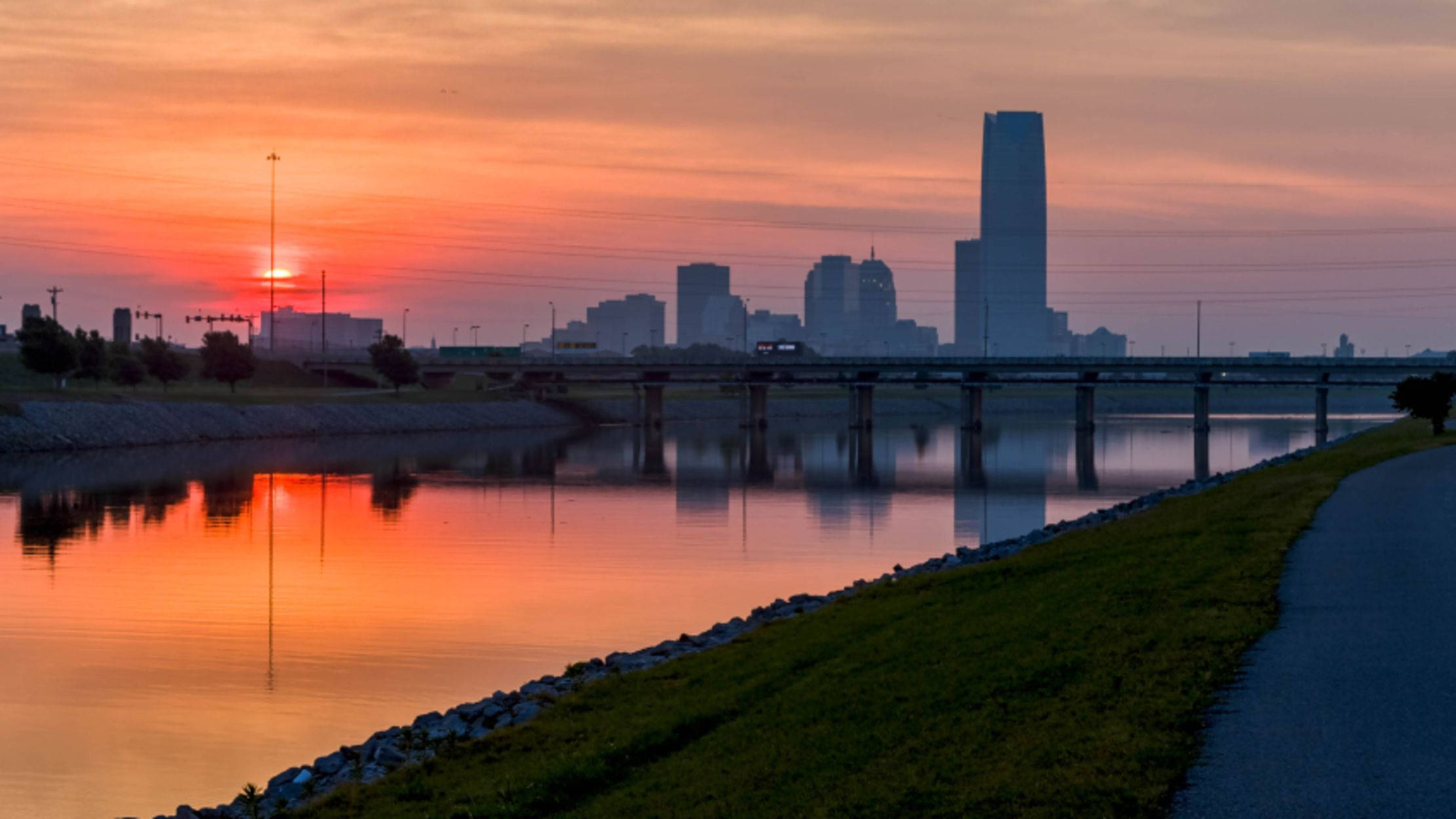 Oklahoma 桌布 壁紙 — 日落時分的城市天際線倒映在平靜的水面上，前景有一座橋樑和一條草地小徑。