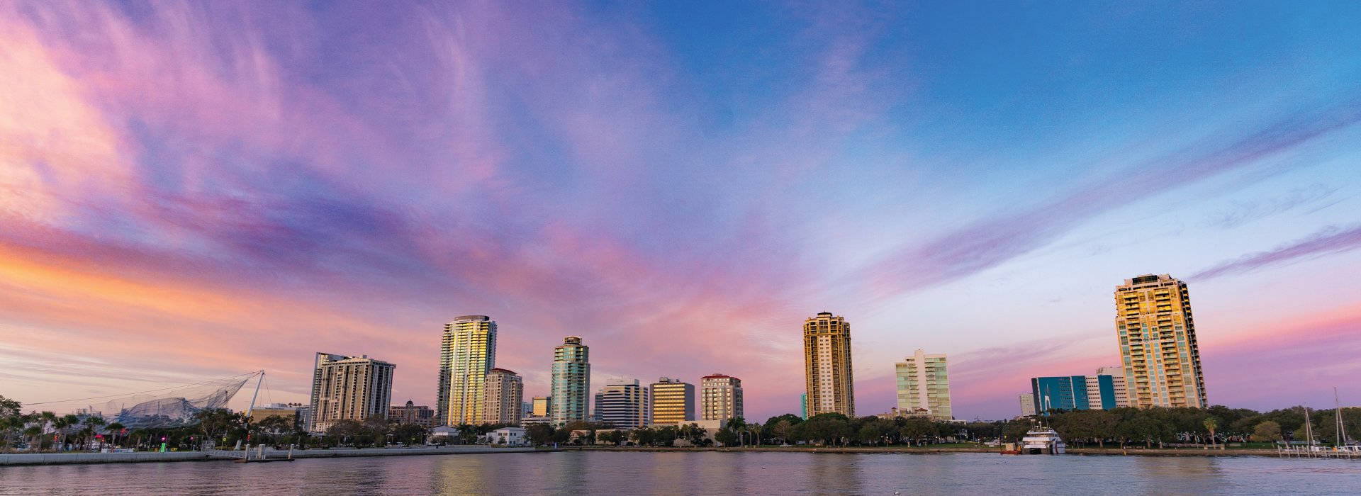 Pink City 桌布 壁紙 — 日落時分的城市天際線全景，充滿活力的粉紅色、紫色和橙色雲彩倒映在平靜的水面上。