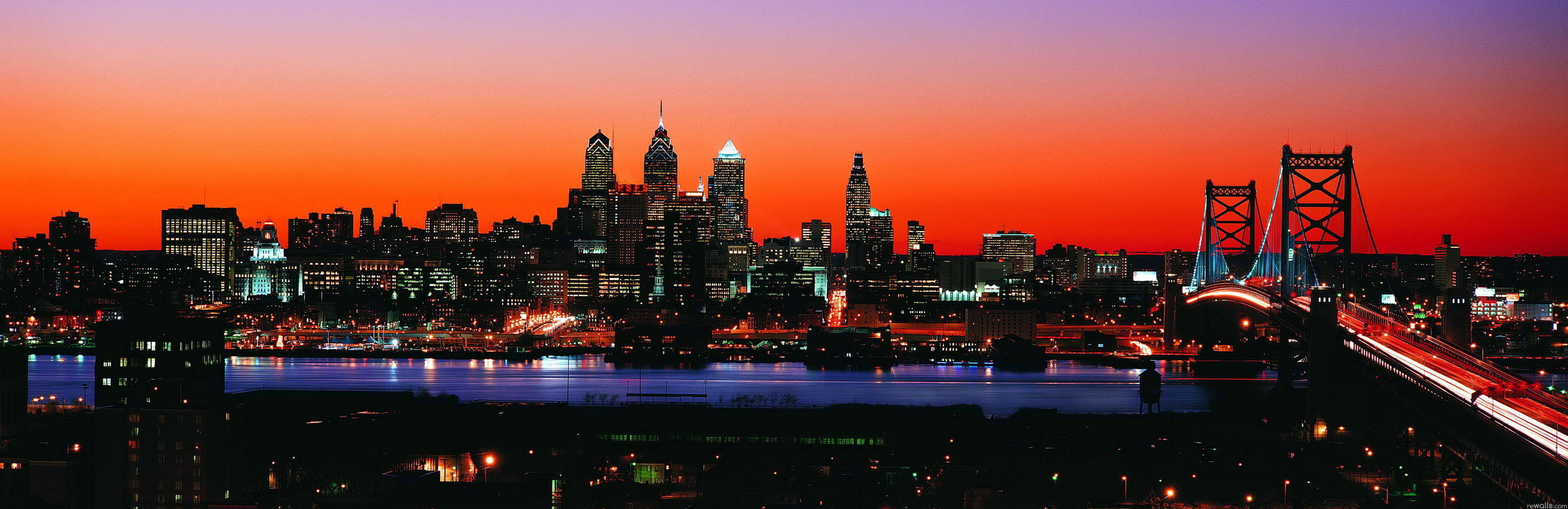 Philadelphia 桌布 壁紙 — 日落時分的城市天際線全景，發光的摩天大樓和一座橋樑在水面上反射出橙色和紫色的色調。
