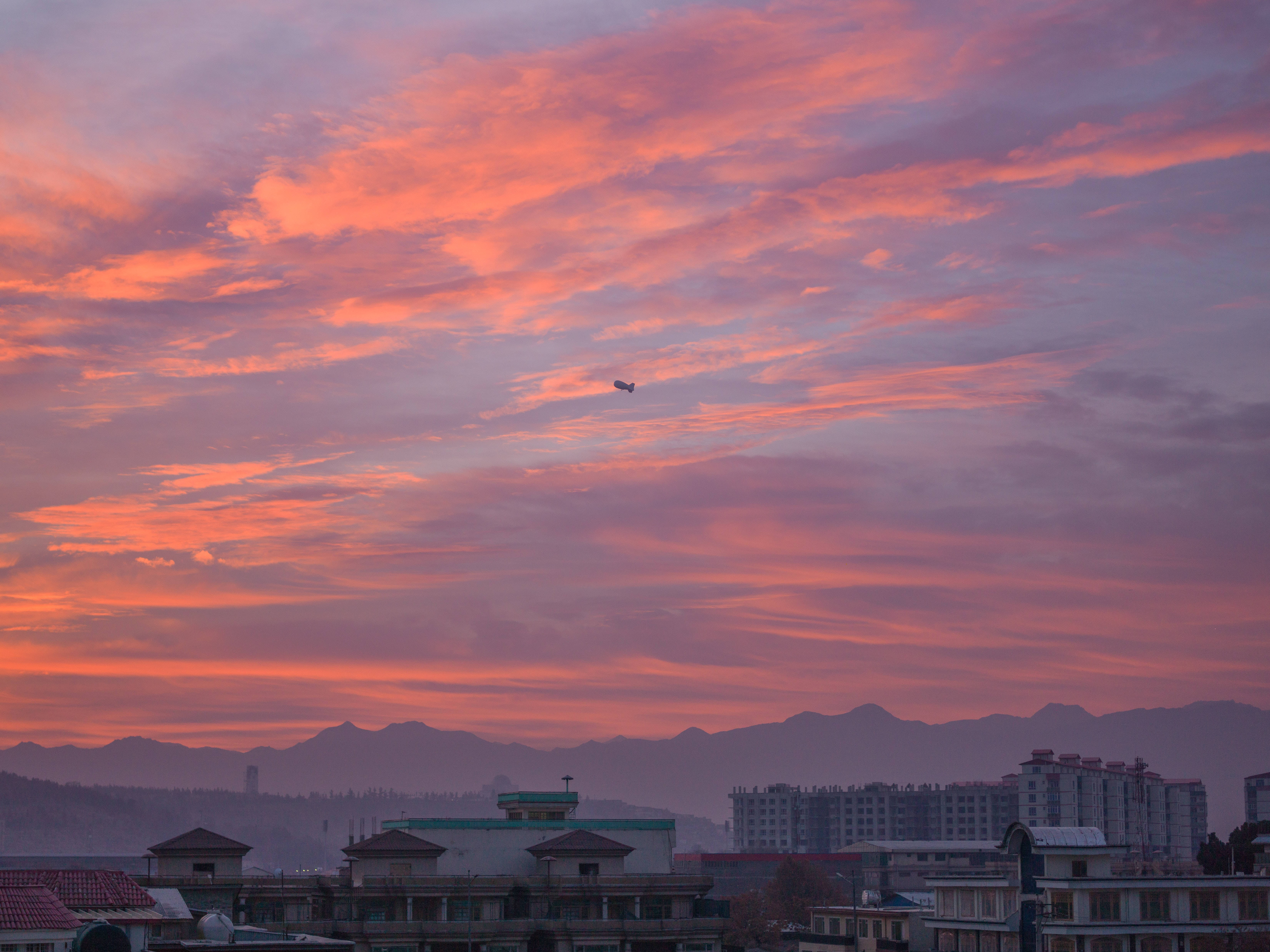 Kabul 桌布 壁紙 — 日落時分的城市景觀，遠山和屋頂上方有火熱的橙色和粉紅色雲彩。