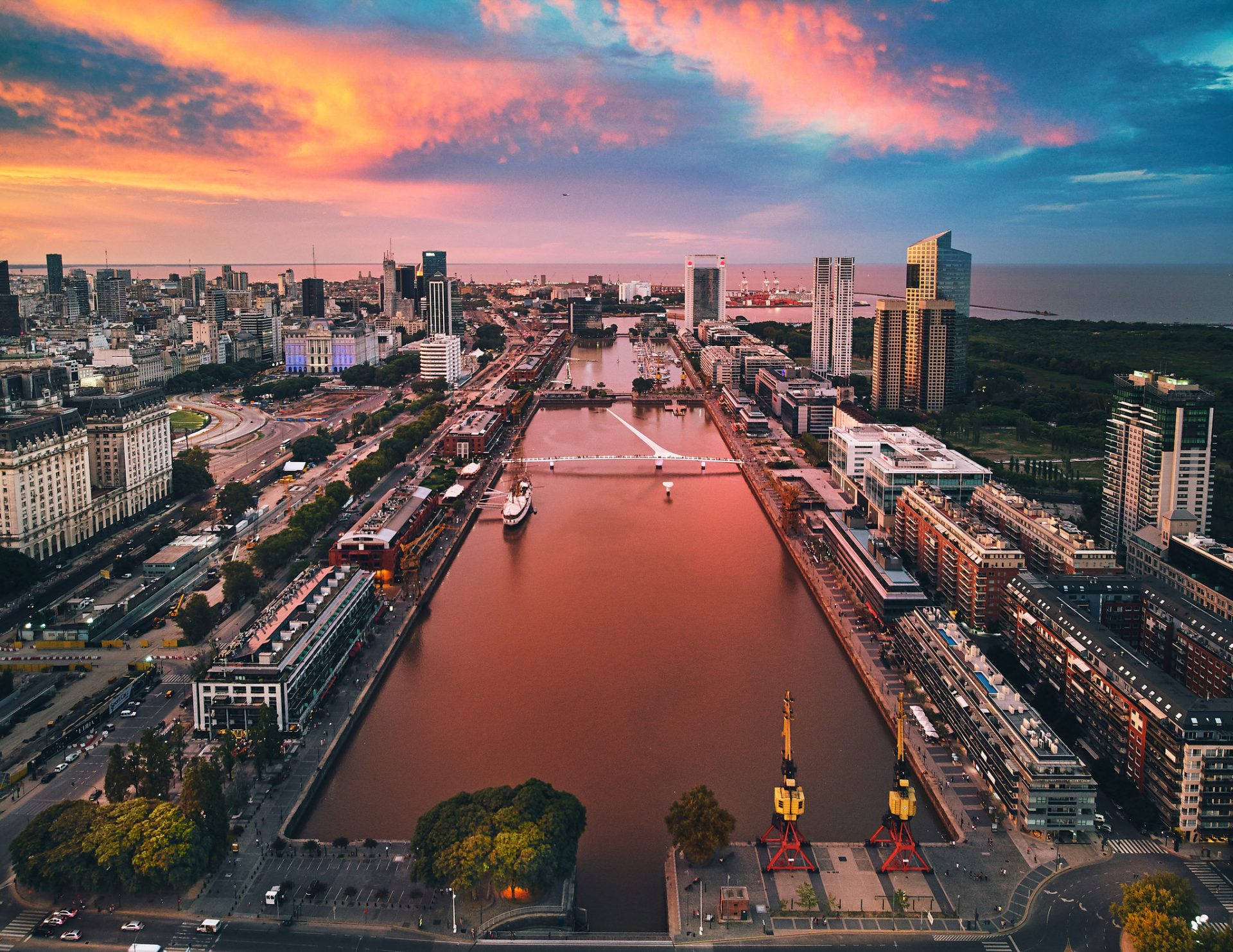 Aires 桌布 壁紙 — 日落時分的城市景觀鳥瞰圖，寬闊的河流倒映著現代和歷史建築上方火熱的橙色和粉紅色雲彩。
