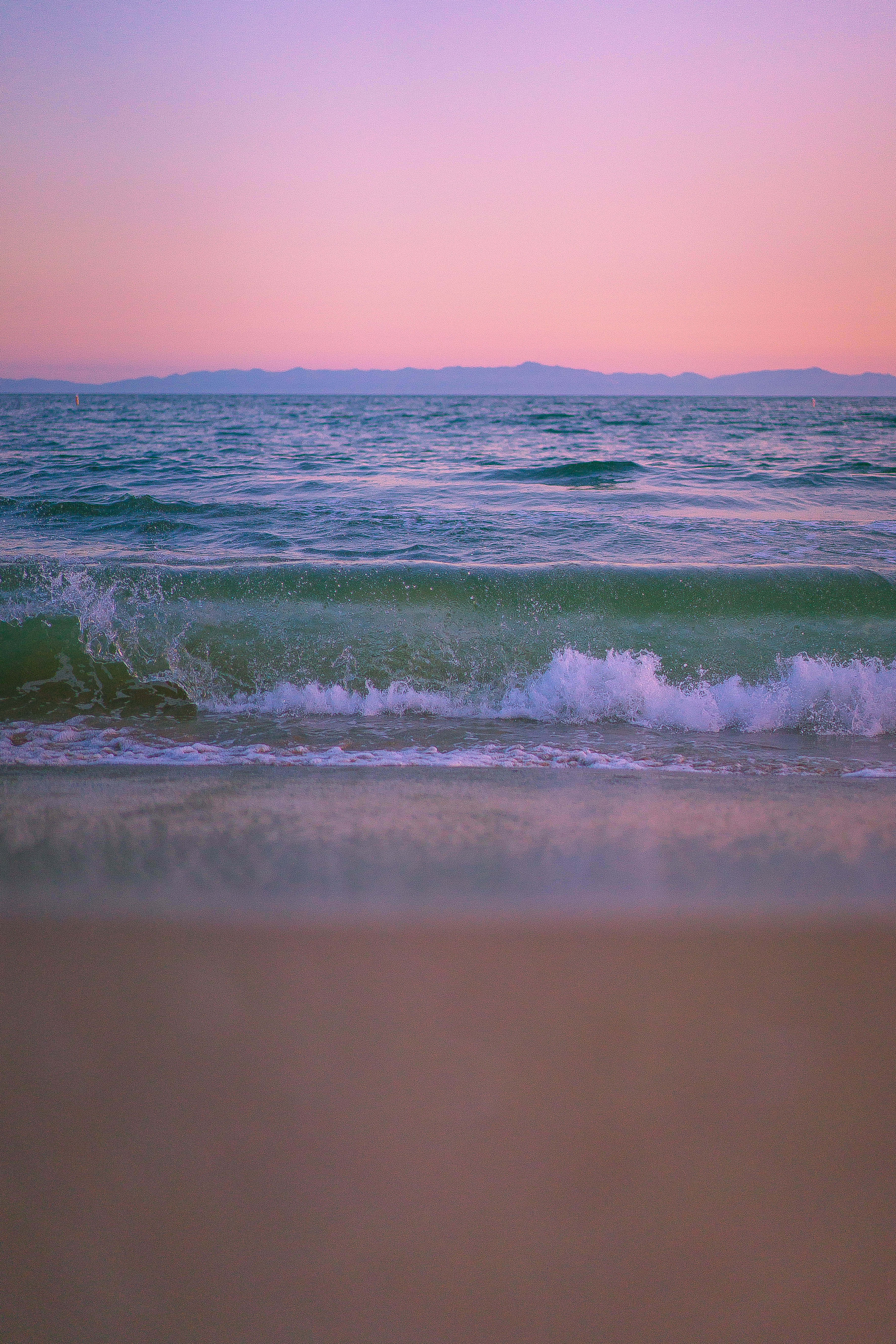 Pink Beach Sunset 桌布 壁紙 — 日落時分的寧靜海灘，粉紅和紫色的天空倒映在碧綠的海浪上，輕輕拍打著柔軟的沙灘。