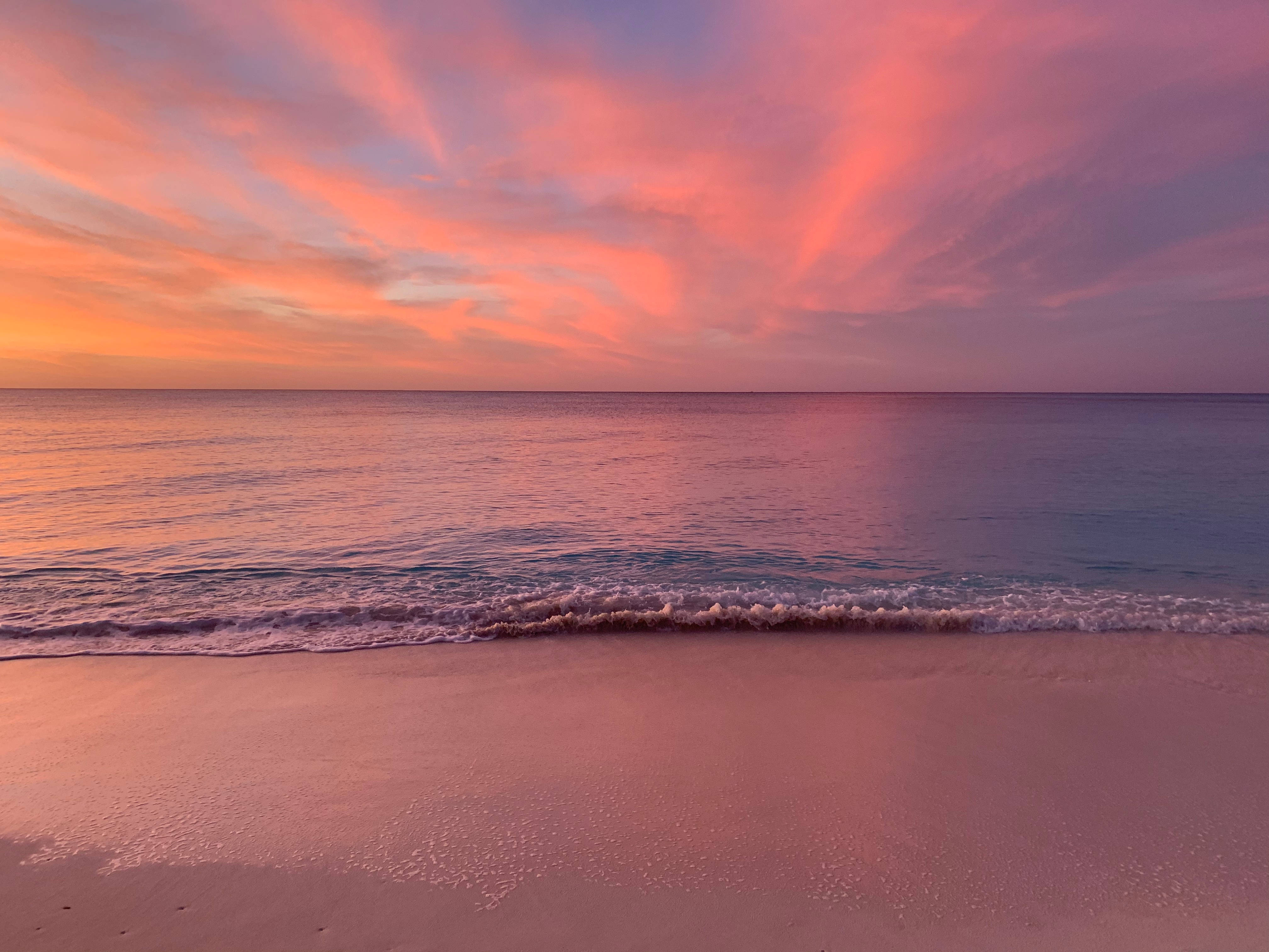 日落 桌布 壁紙 — 日落時分的寧靜海灘，鮮豔的粉紅、橙色和紫色雲彩倒映在平靜的碧綠海水中。