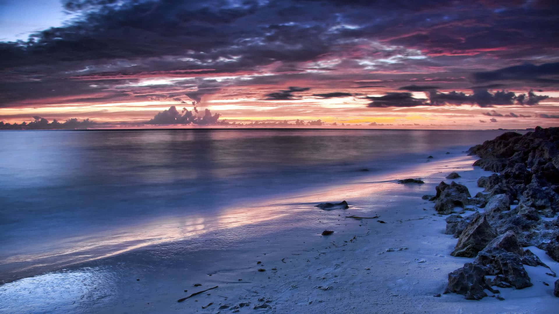 Beach At Night 桌布 壁紙 — 日落時分的寧靜海灘，鮮豔的紫色和橙色雲彩倒映在岩石海岸旁的平靜水面上。