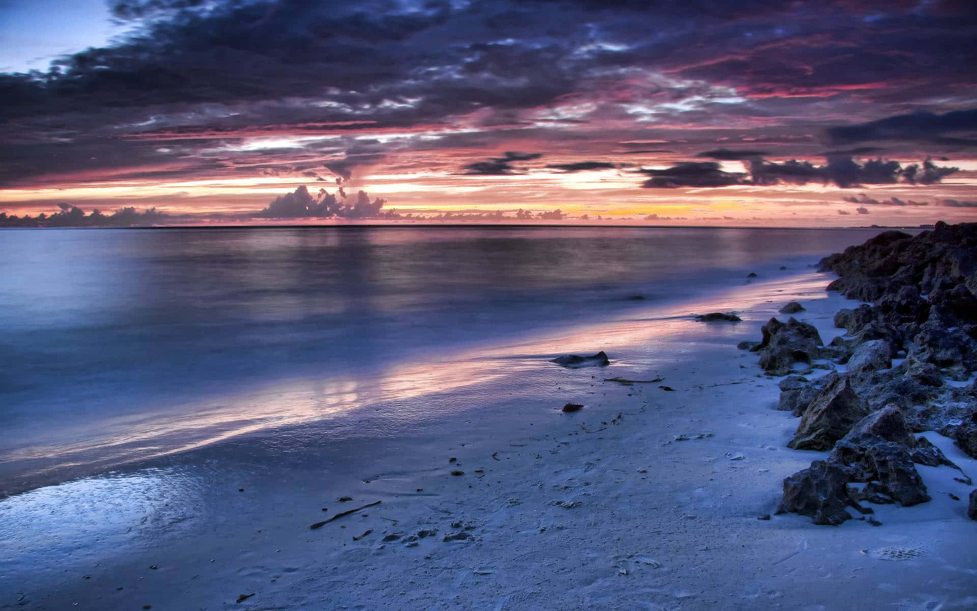 Scenic Desktop 桌布 壁紙 — 日落時分的寧靜海灘，鮮豔的紫色和橙色雲彩倒映在岩石海岸旁的平靜水面上。